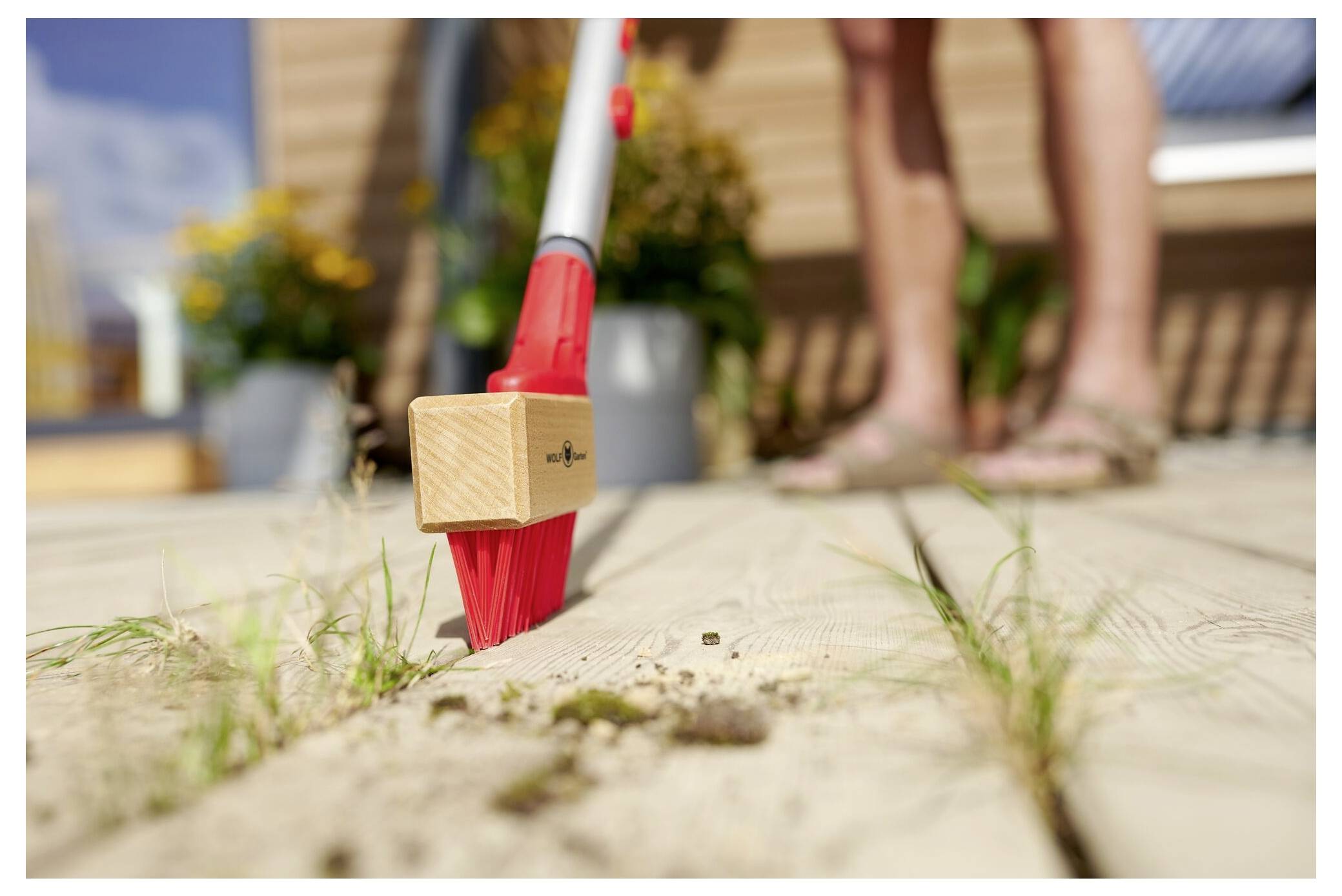 Una persona usa una scopa rossa per spazzare sporco e piccoli detriti da un ponte di legno con piante sullo sfondo, suggerendo una pulizia esterna.