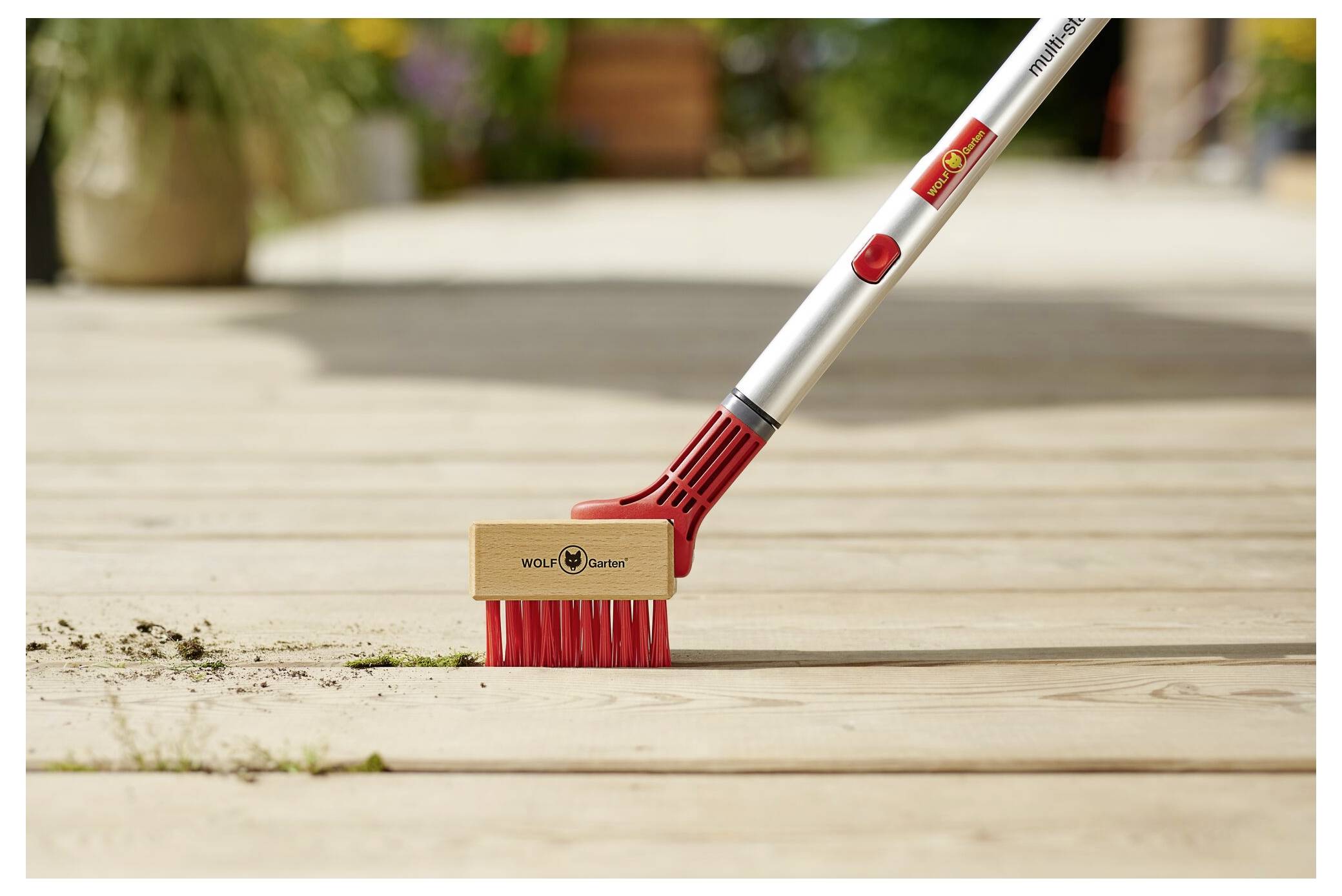 Un primo piano di una spazzola rossa e argentata con una testa in legno, che rimuove sporco e erbacce dagli spazi vuoti di un ponte in legno. La spazzola ha un logo del marchio.