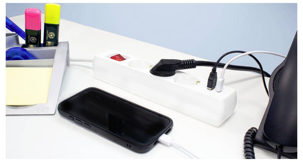 Uno smartphone in carica da una presa multipla su una scrivania, insieme a forniture da ufficio tra cui evidenziatori, nastro adesivo e foglietti adesivi.