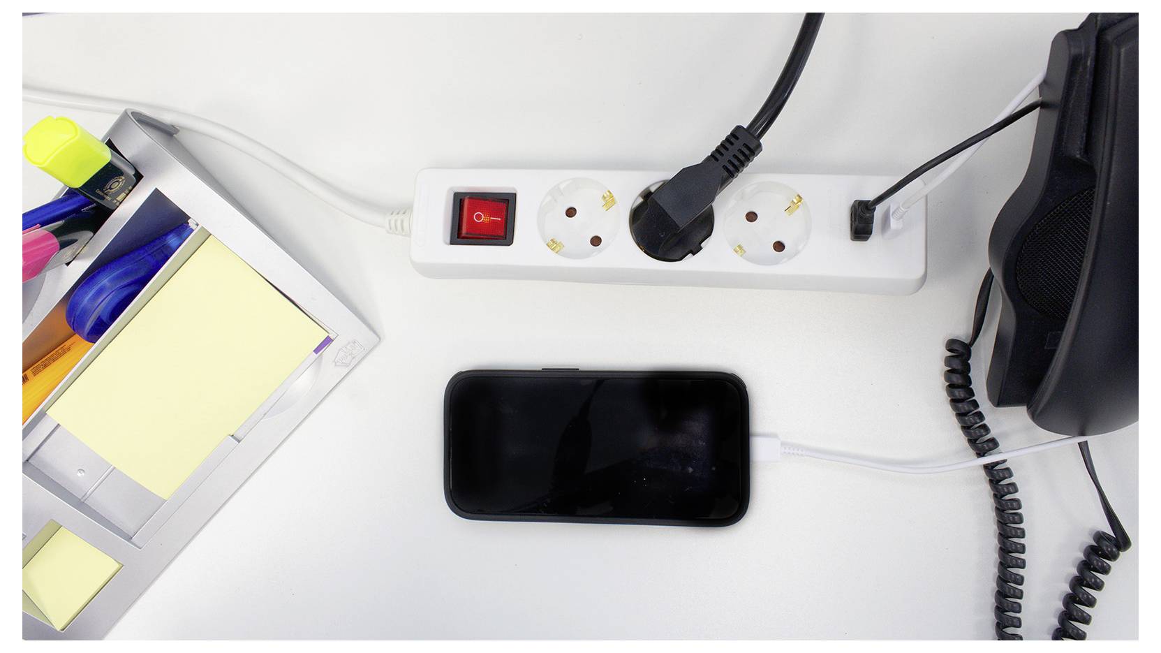 Uno smartphone è in carica su una scrivania, collegato a una ciabatta con un interruttore rosso. Nelle vicinanze ci sono materiali per ufficio e un telefono.