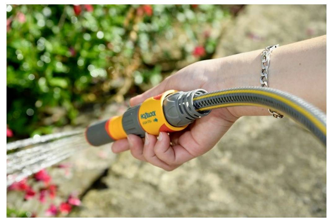 Una persona che innaffia le piante con un tubo da giardino, circondata da fiori in una giornata soleggiata.