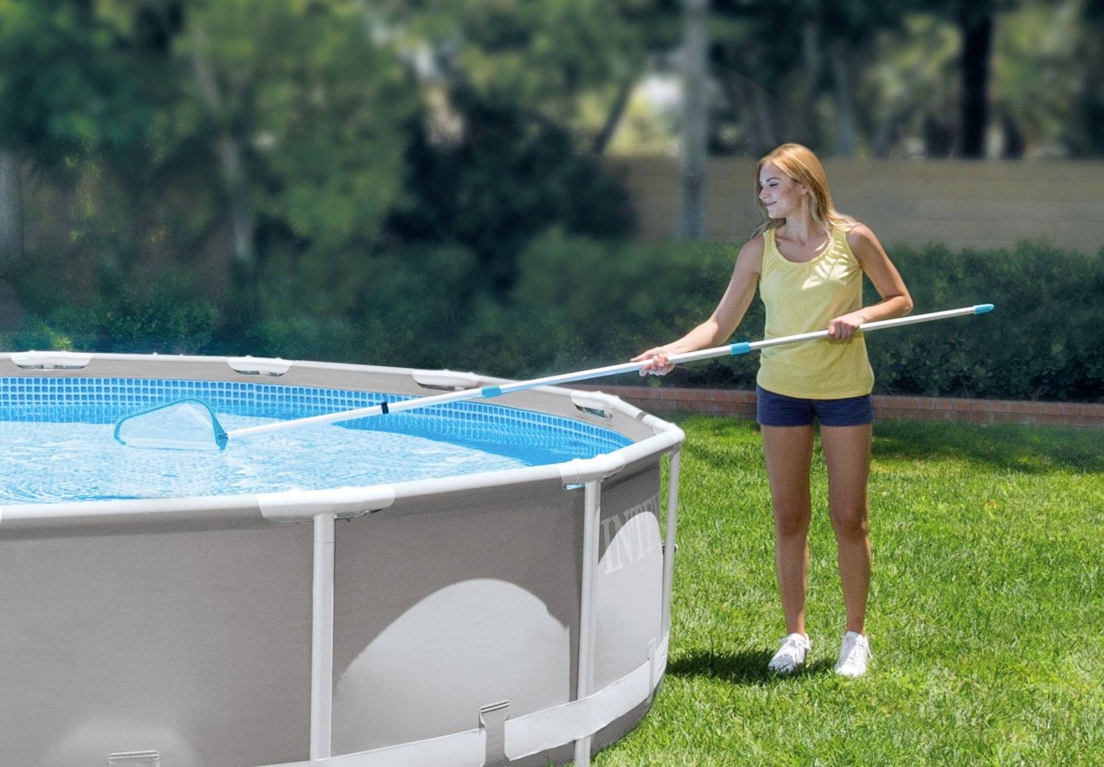 Una donna in una canotta gialla e pantaloncini pulisce una grande piscina rotonda all'aperto con un retino per sfiorare in un giardino erboso.