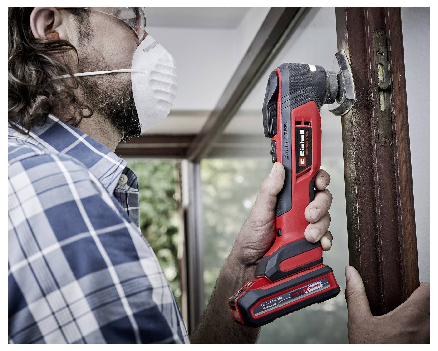 Una persona che indossa occhiali di sicurezza e una mascherina utilizza un attrezzo elettrico rosso per lavorare su una struttura in legno accanto a una finestra.