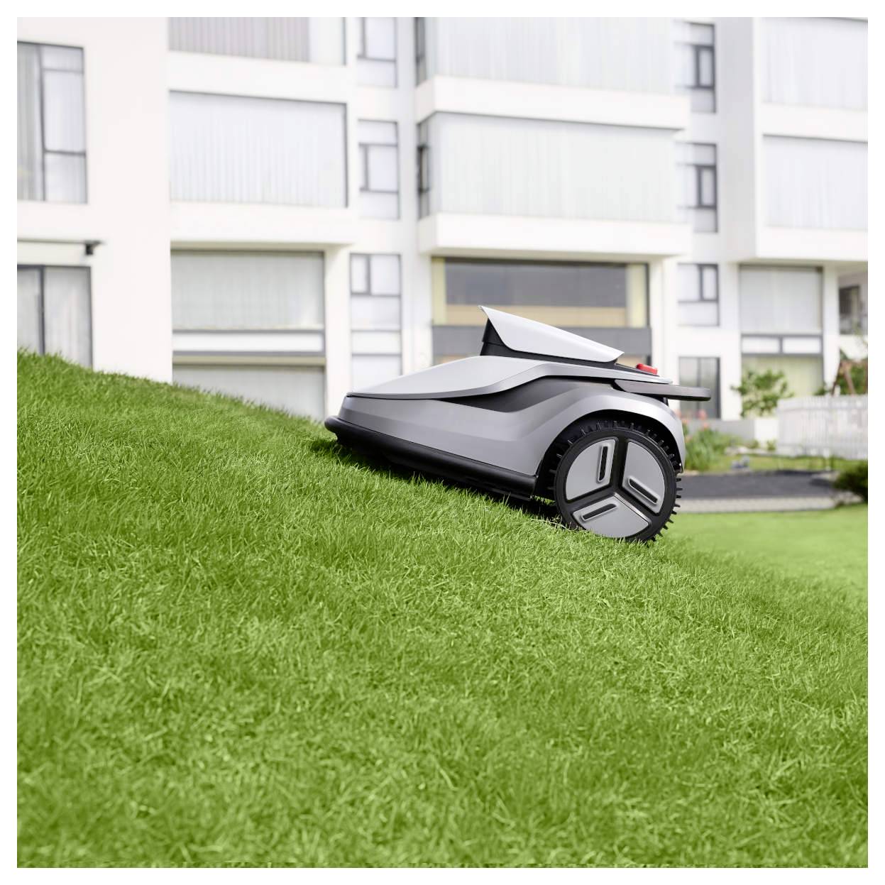 Un tagliaerba robotico in funzione, che attraversa agevolmente un prato in pendenza, con moderni edifici residenziali sullo sfondo.