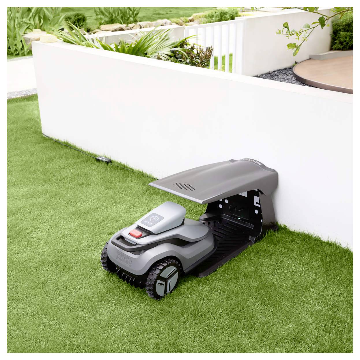Un tosaerba robotico è parcheggiato sotto un piccolo rifugio accanto a un muro bianco in un giardino, mostrando il suo design compatto e la soluzione di archiviazione.