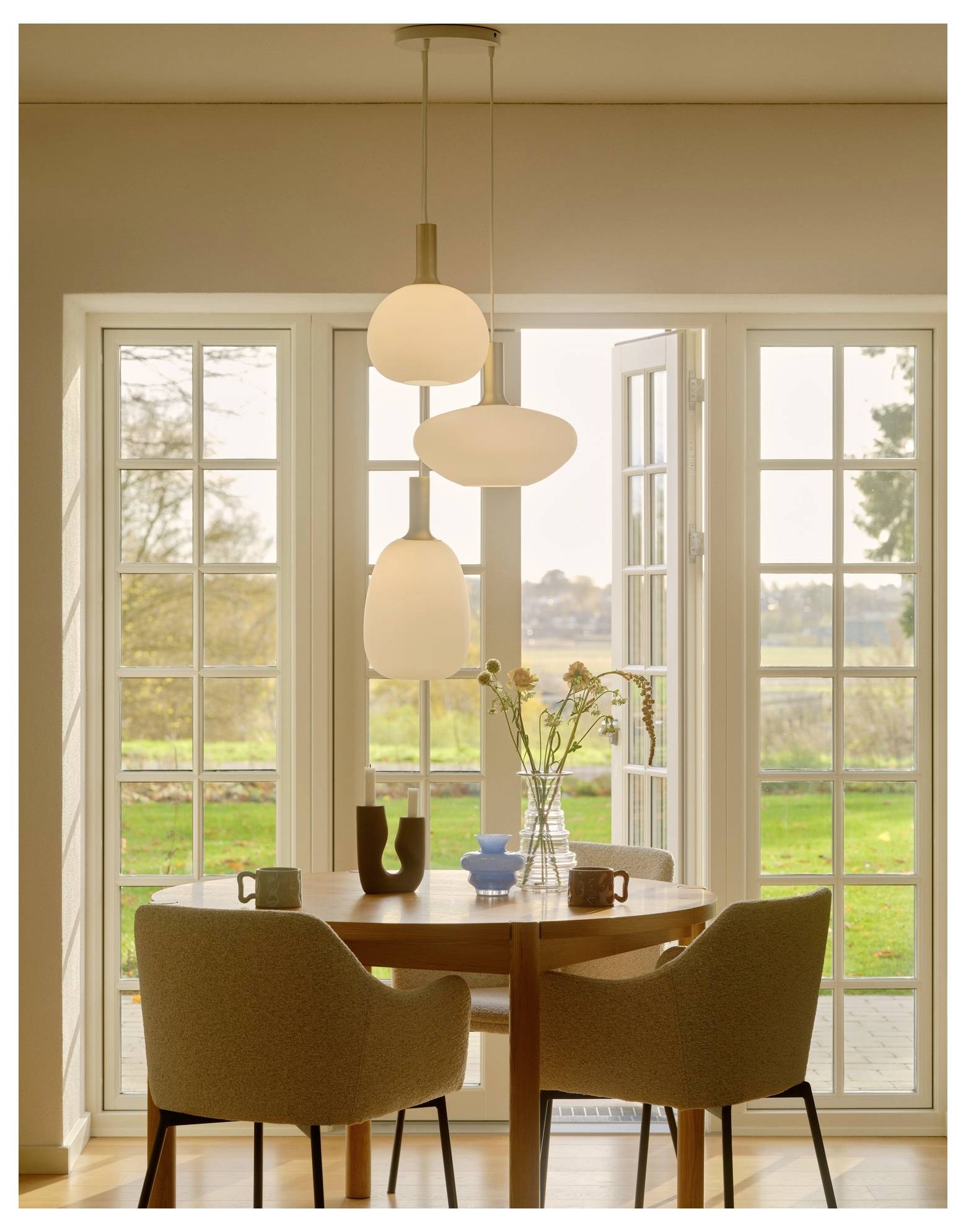 Un'accogliente zona pranzo con un tavolo rotondo, due sedie beige, lampadari a sospensione moderni e una vista dalla finestra di un giardino verde all'esterno.