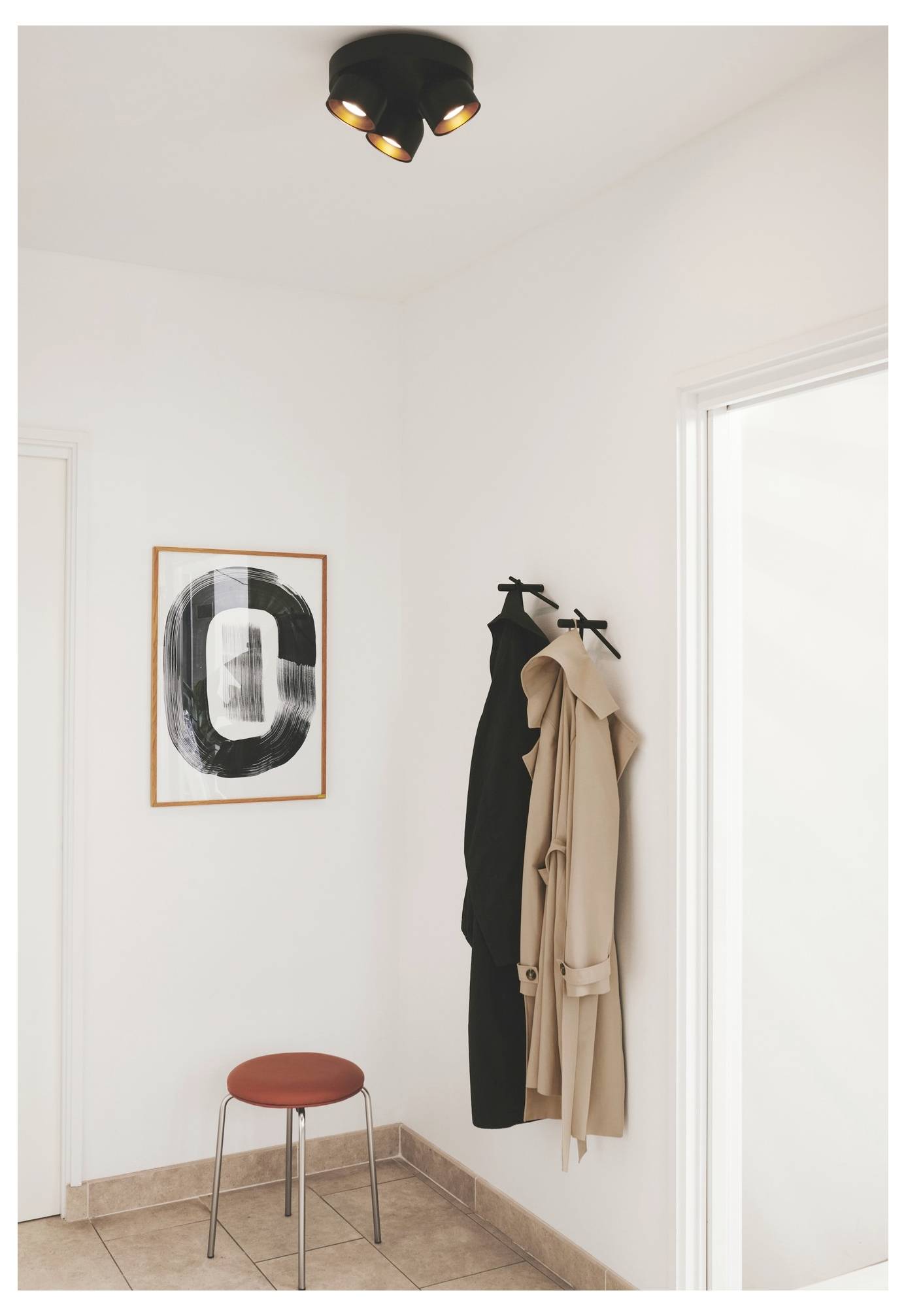Un angolo minimalista di un corridoio con uno sgabello, due cappotti appesi e un'opera d'arte astratta sulla parete. Una lampada a soffitto nera sopra.