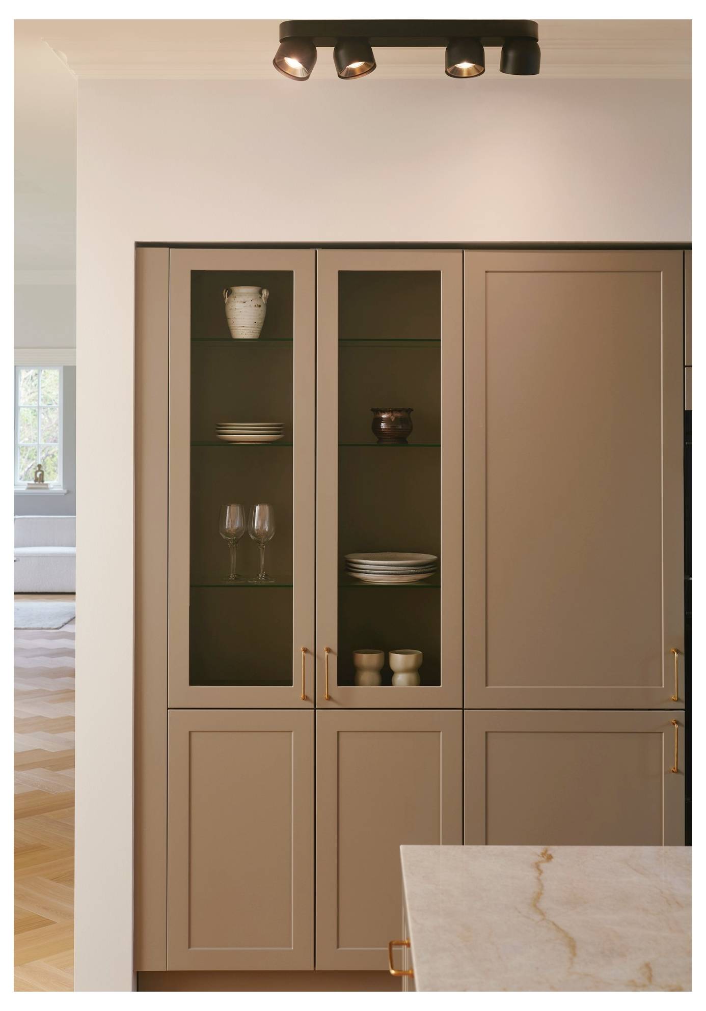Una cucina moderna con armadietti beige con porte in vetro che mostrano piatti e bicchieri, sotto un'illuminazione a binario al soffitto.