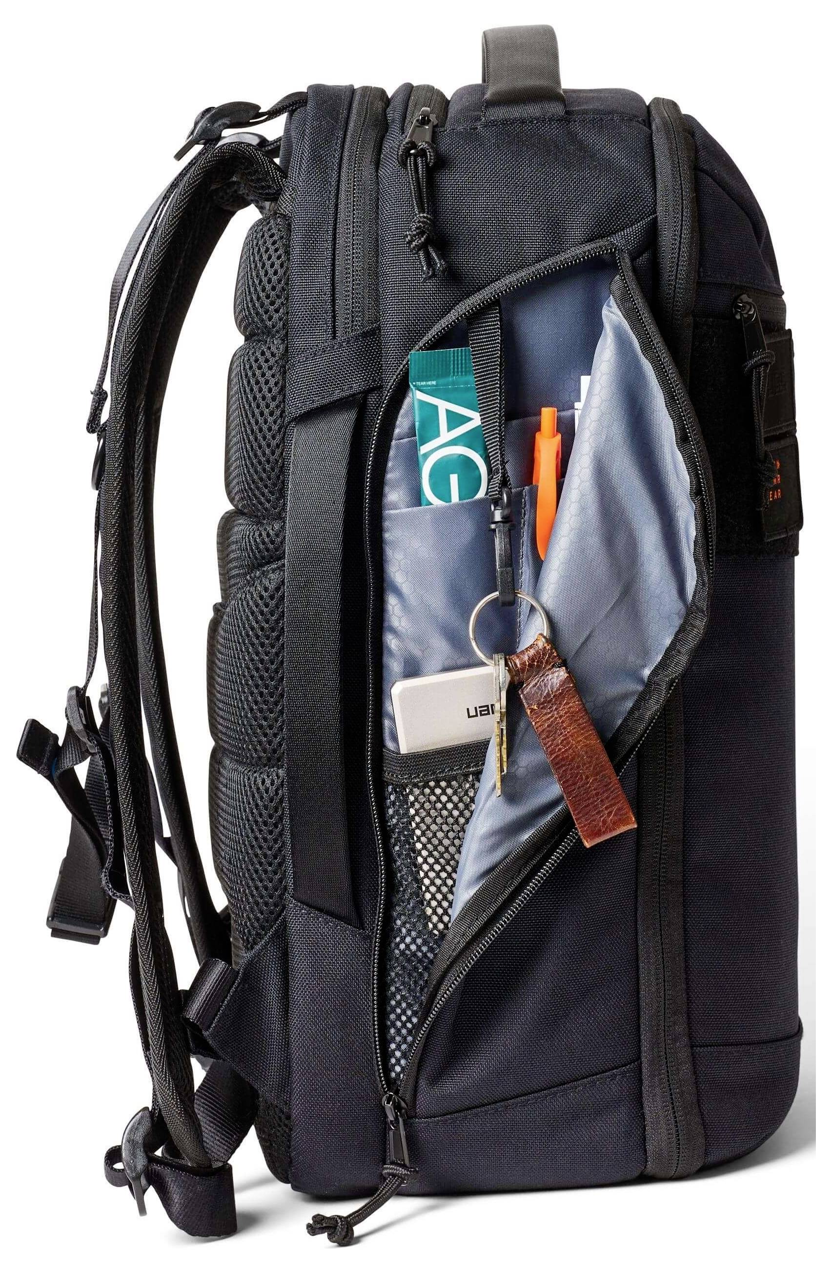 Uno zaino nero aperto davanti, che rivela scomparti organizzati con un portachiavi, una penna, una rivista e un piccolo dispositivo all'interno.