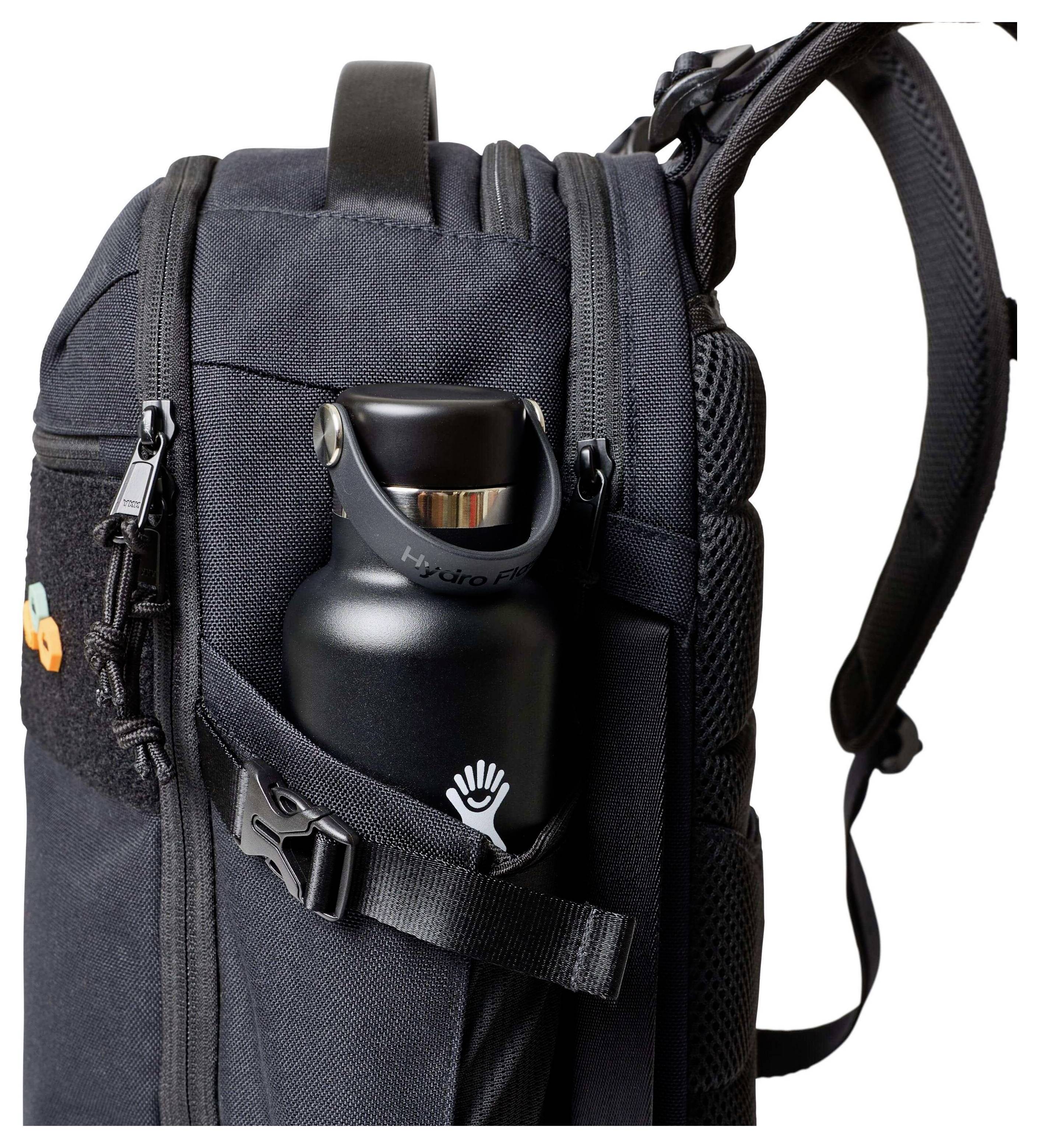 Uno zaino nero con più scomparti, che mostra una bottiglia d'acqua infilata in una tasca laterale con una cinghia che la fissa.