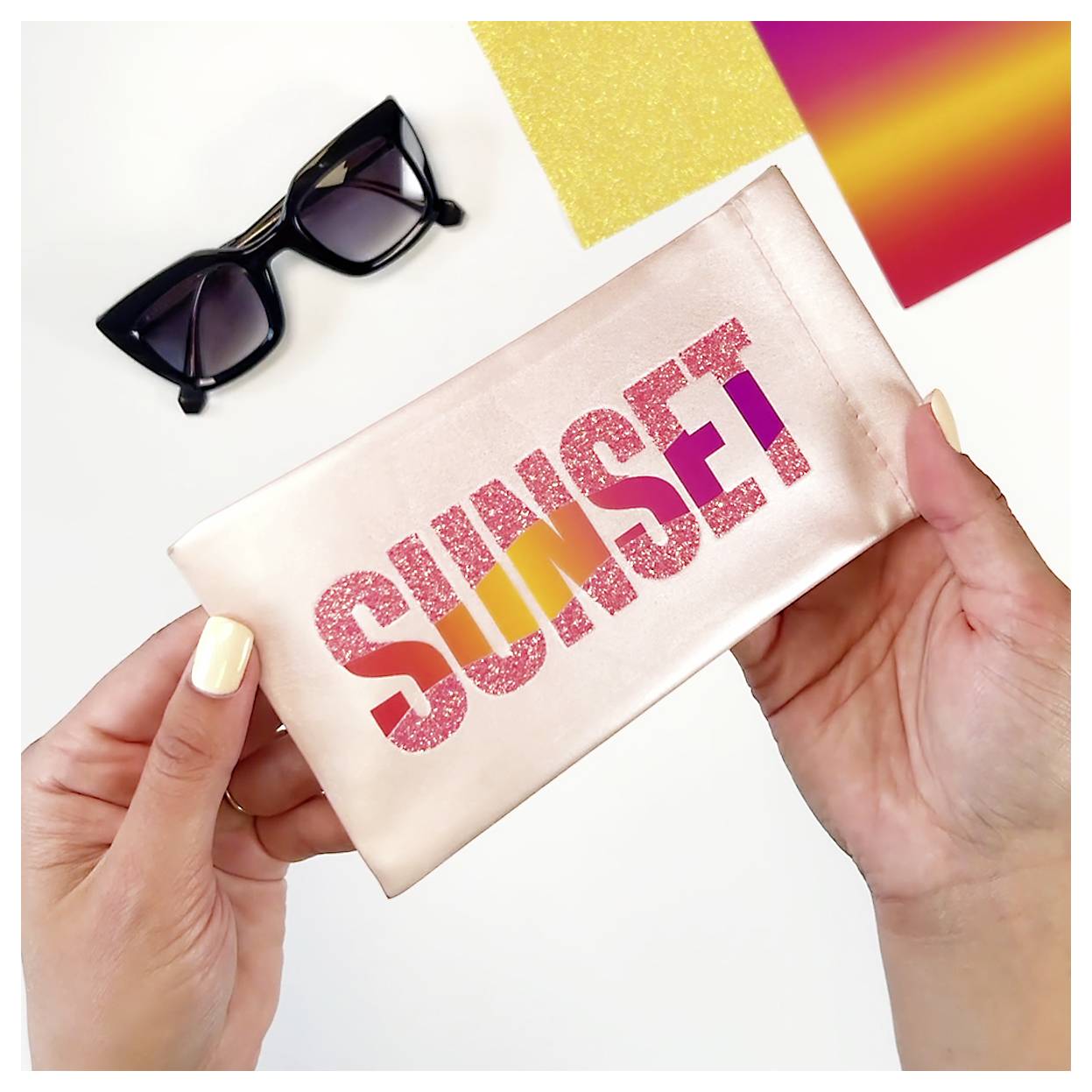 Le mani tengono una borsa lucida etichettata 'SUNSET' con lettere glitterate; nelle vicinanze ci sono occhiali da sole e campioni di colori vibranti.