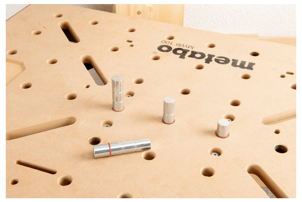 Un piano di lavoro in legno con vari fori e quattro pioli metallici cilindrici inseriti. Il logo 'metabo' e il modello 'MFT K 700' visibili.