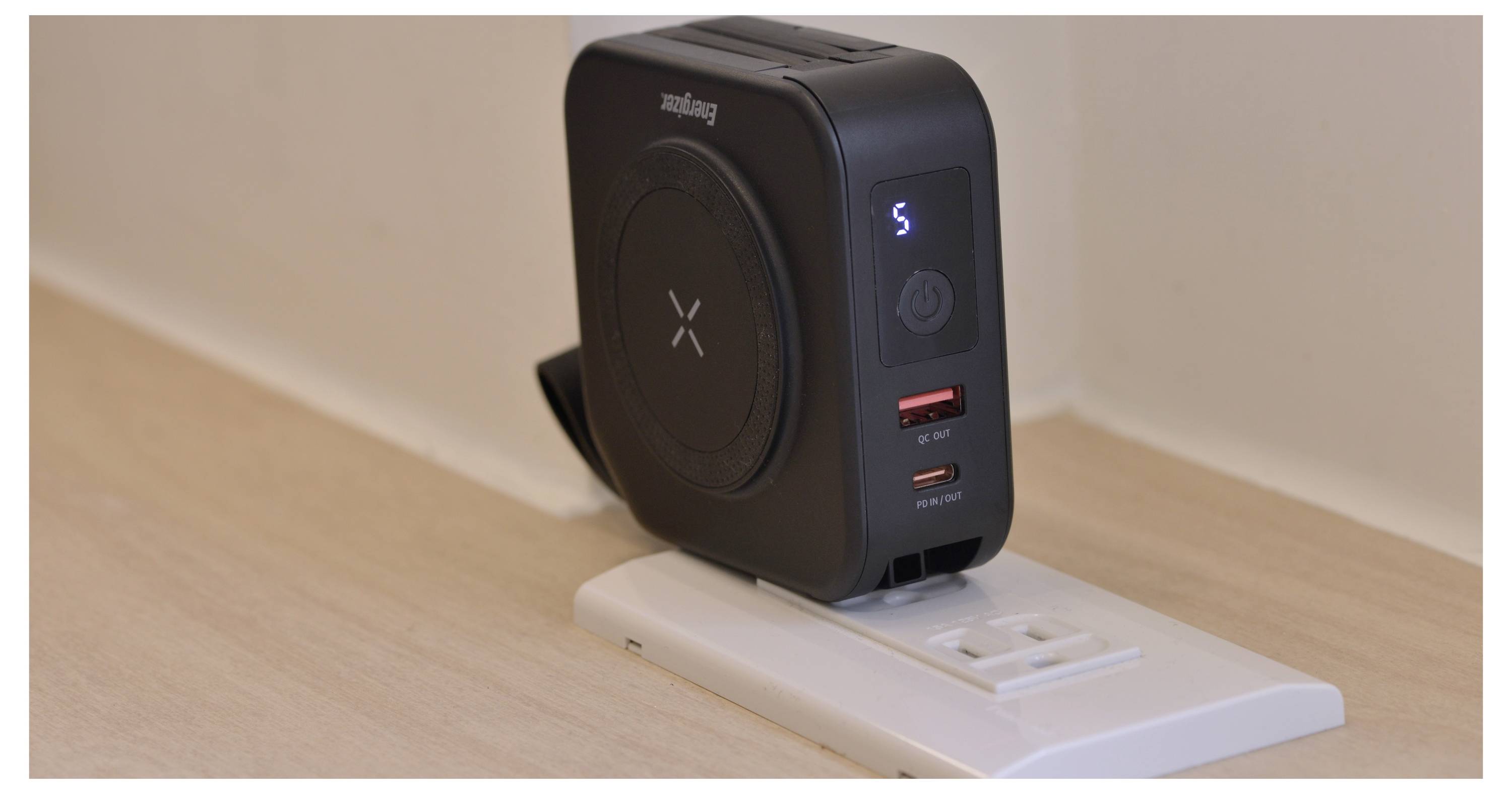 Un caricabatterie wireless nero e un adattatore di alimentazione collegati a una presa a muro, con indicatori LED e due porte USB su una superficie di legno chiaro.