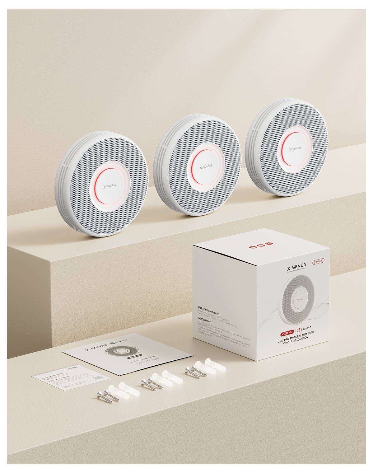 Tre rilevatori di fumo circolari sono disposti in fila su una superficie beige. Davanti, un set in scatola include manuali e accessori di montaggio.