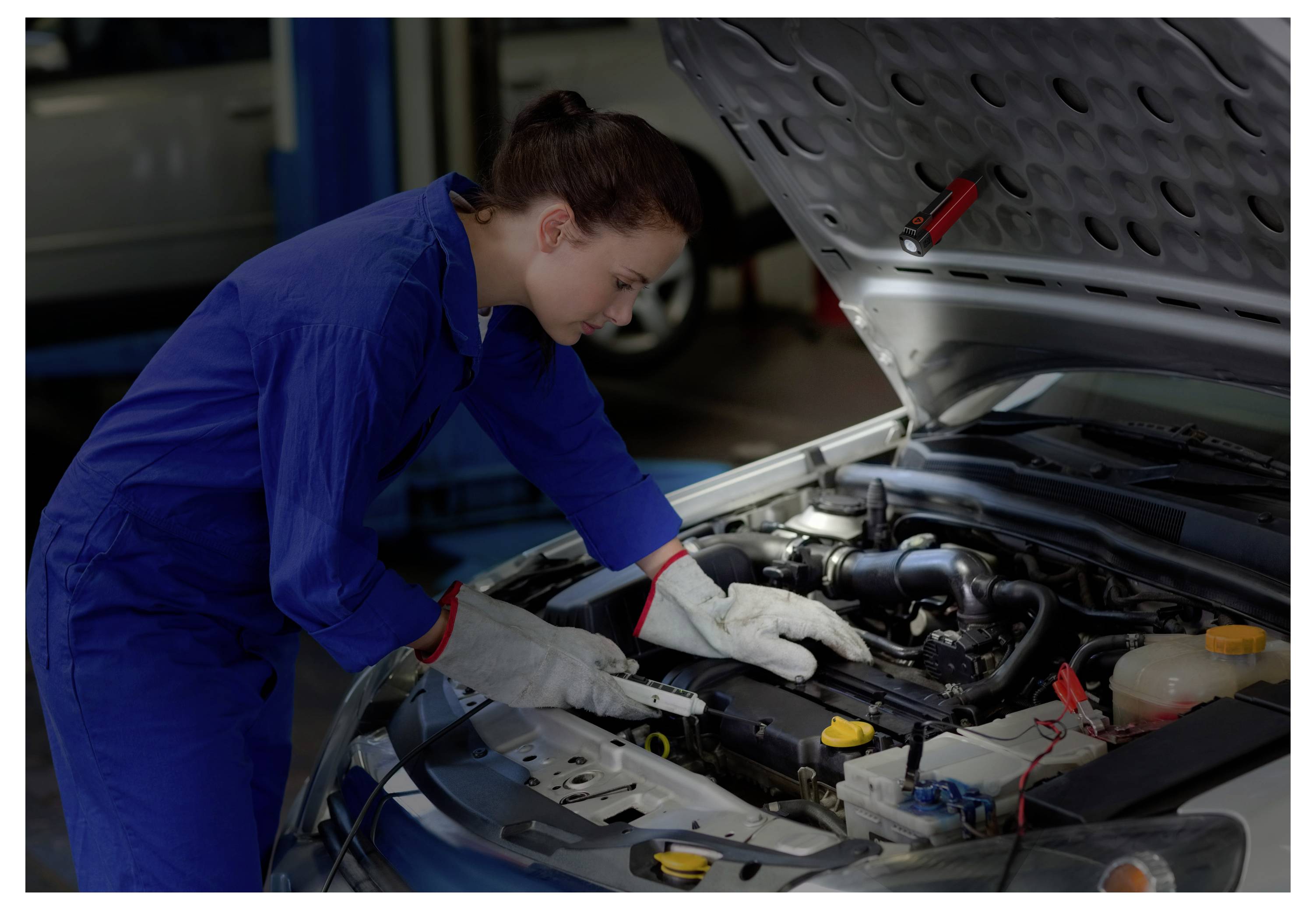 Un meccanico in tuta blu ispeziona il motore di un'auto sotto il cofano aperto in un'officina di riparazione auto.