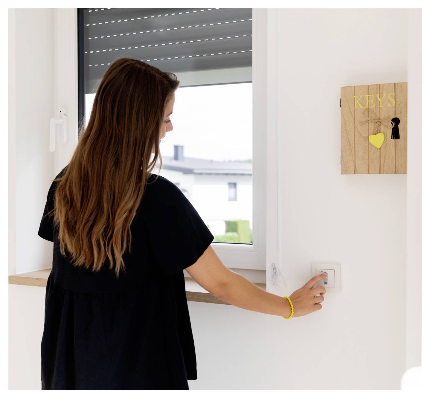 Una donna in una maglietta nera preme un pulsante su un interruttore a parete sotto una finestra. Vicino c'è un portachiavi in legno etichettato 'KEYS' con un buco della serratura.