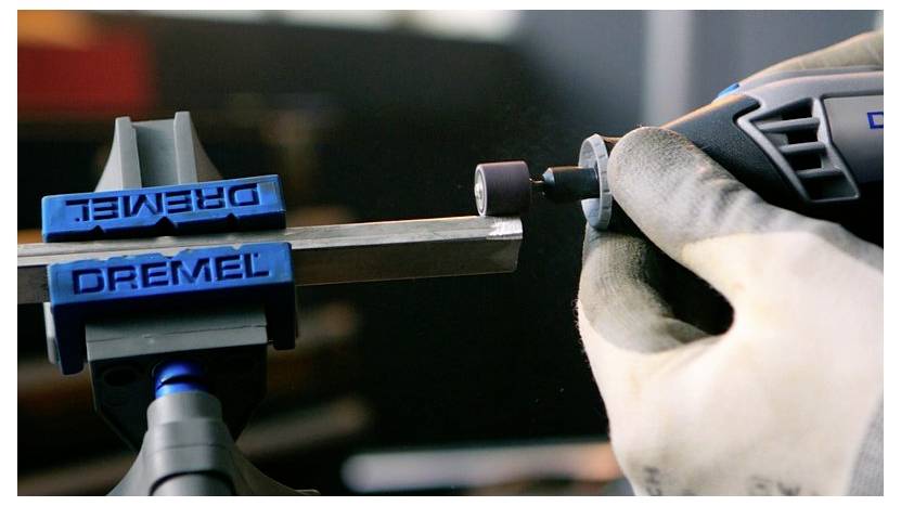 Un primo piano di una persona che utilizza un utensile rotante per levigare il bordo di una barra metallica bloccata in una morsa blu, mostrando la precisione dello strumento.