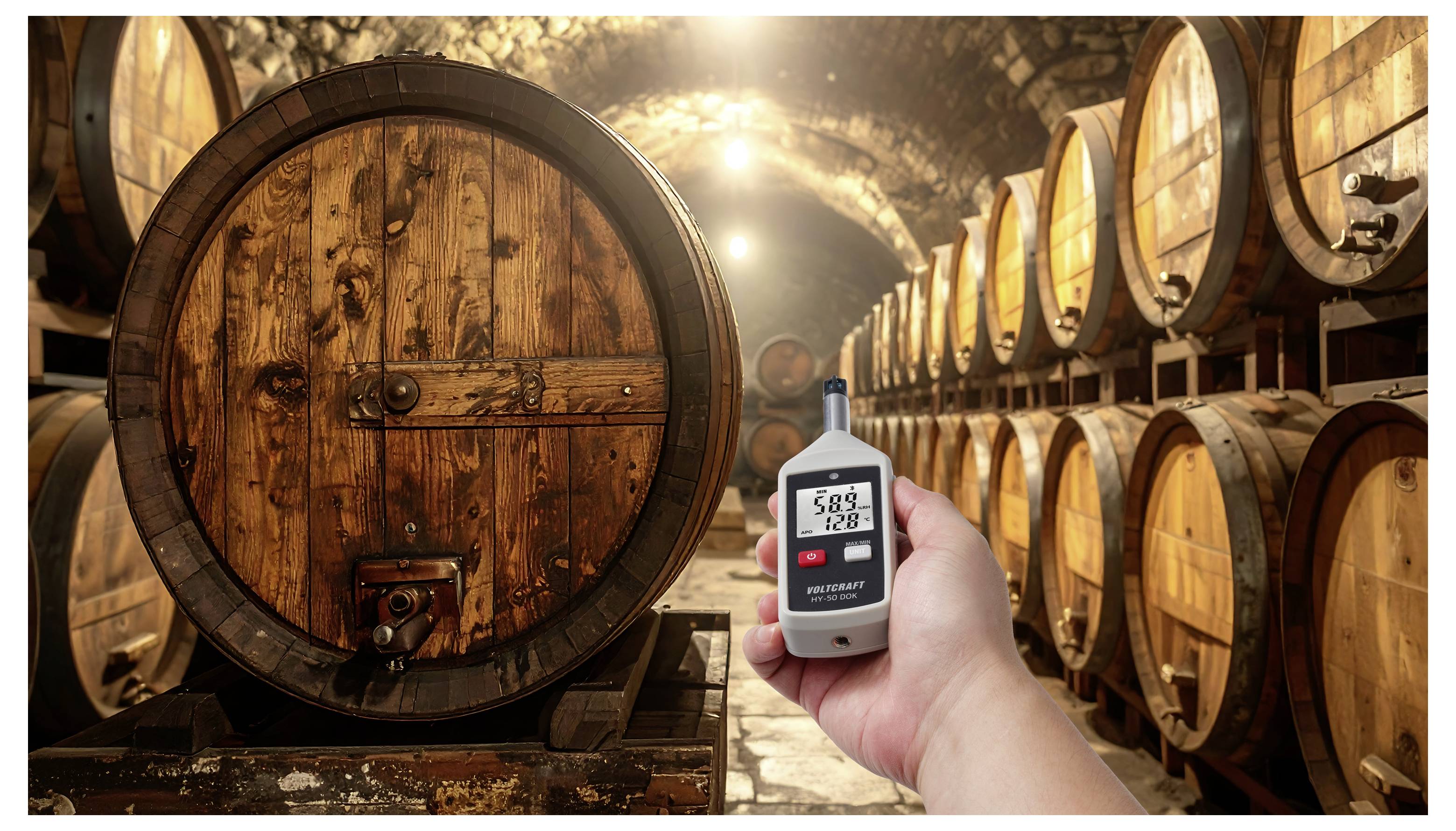 Mano che tiene un misuratore digitale che mostra 58,8 e 12,8 in una cantina vinicola con botti di legno in fila, suggerendo il monitoraggio della temperatura e dell'umidità.