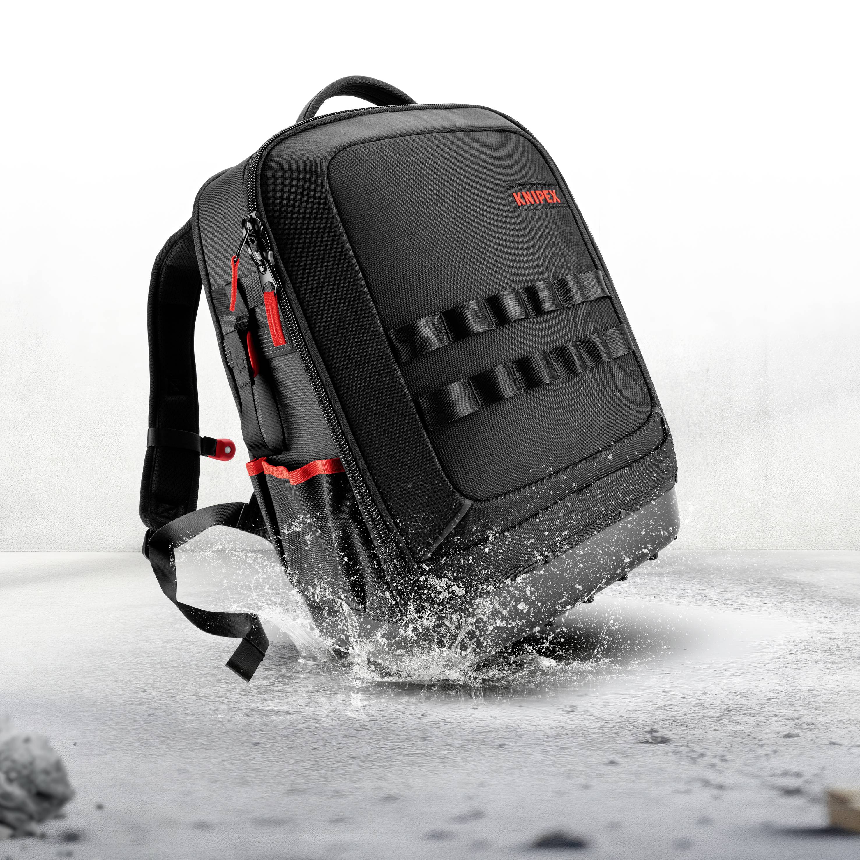 Zaino KNIPR nero con accenti rossi che spruzza attraverso l'acqua, mostrato in posizione verticale su una superficie bianca.