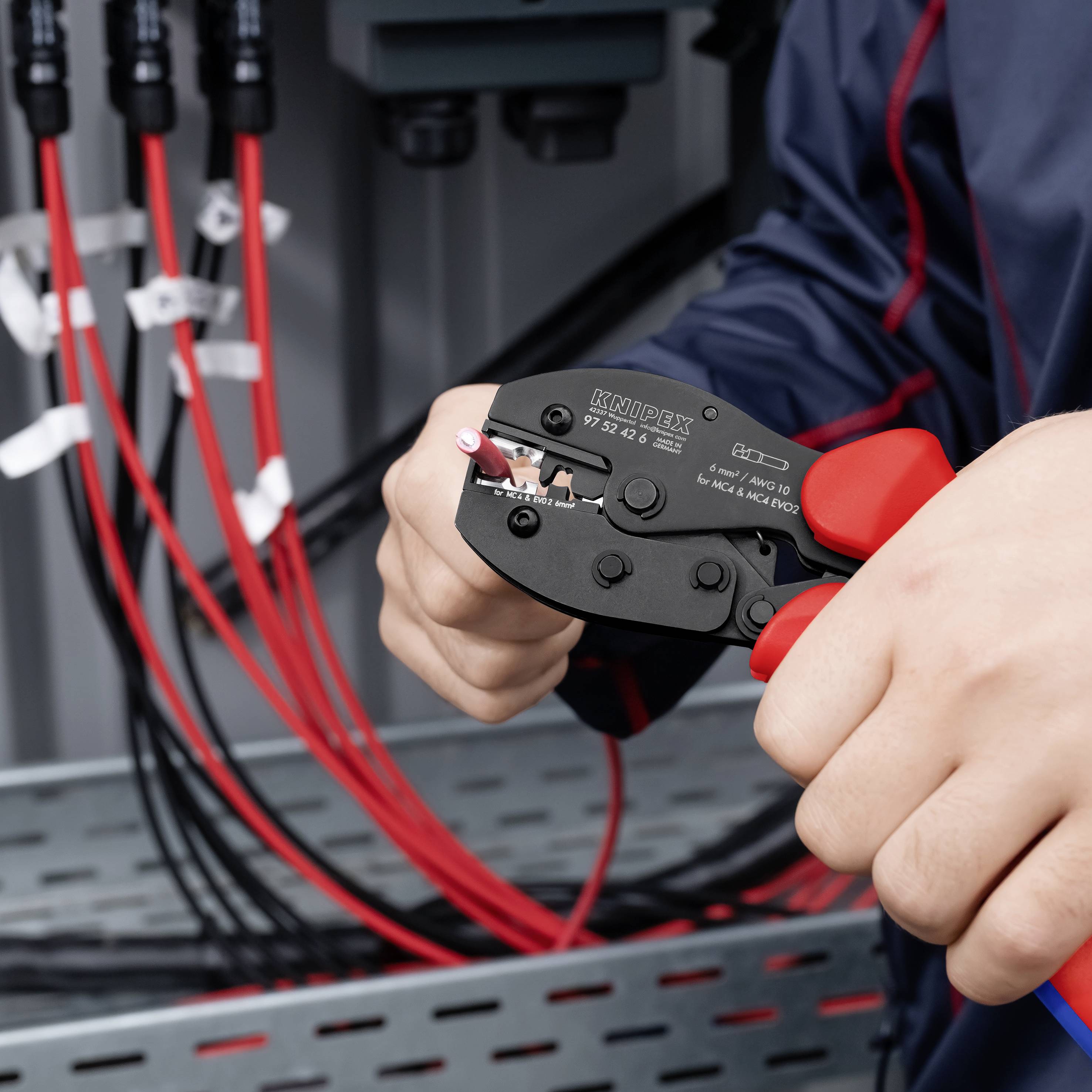 Mani che utilizzano uno spelacavi per spellare un cavo rosso all'interno di un quadro elettrico, con altri cavi rossi e neri visibili sullo sfondo.