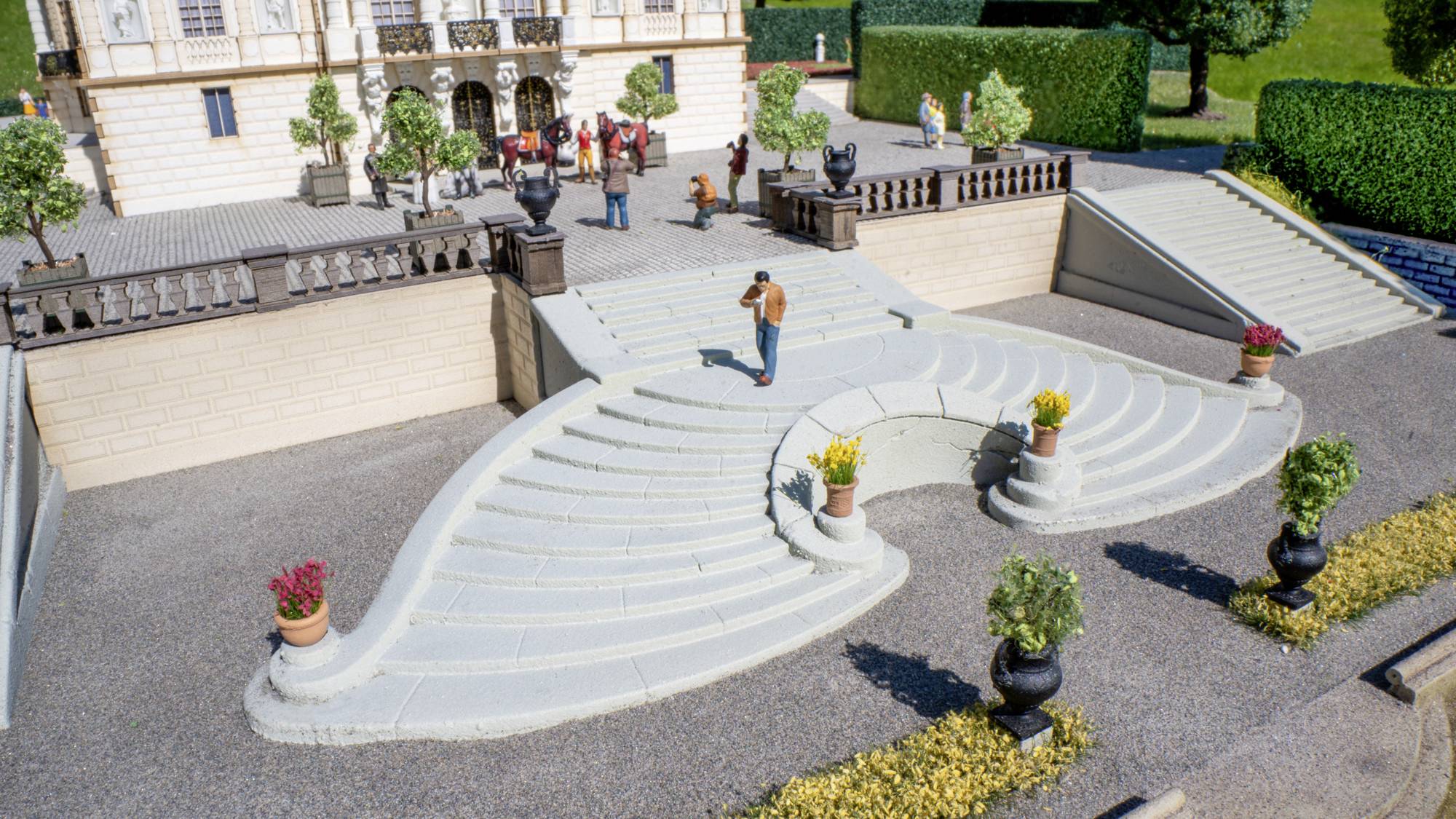 Modello in miniatura di terrazza esterna con scalinate bianche curve che si estendono ampiamente, vasi di fiori e piccole figure davanti a un grande edificio.