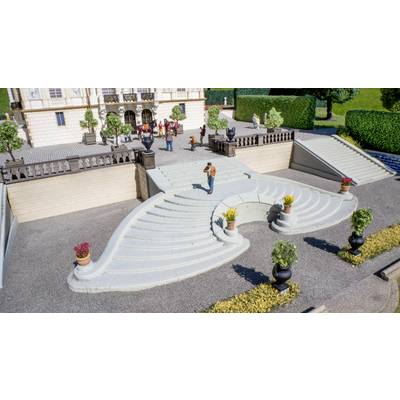 Modello in miniatura di terrazza esterna con scalinate bianche curve che si estendono ampiamente, vasi di fiori e piccole figure davanti a un grande edificio.