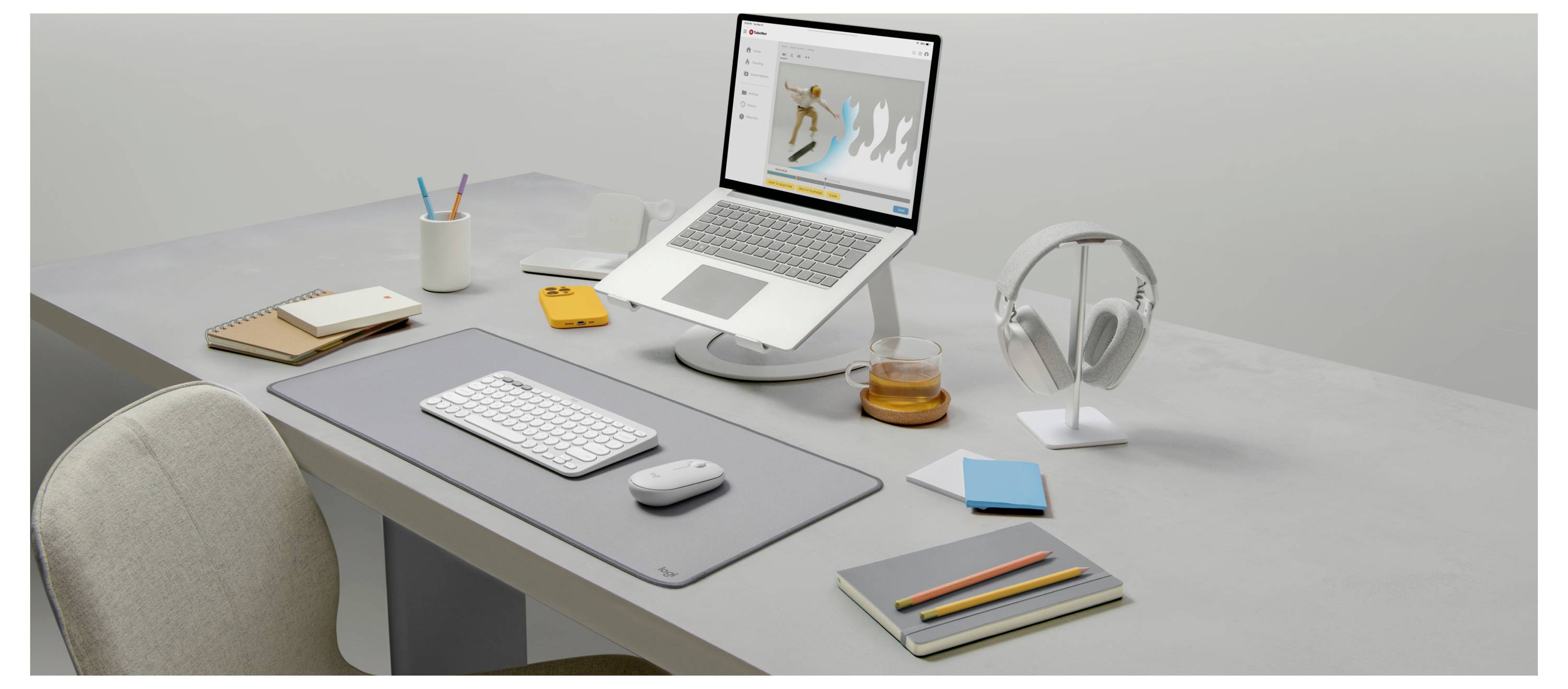 Area di lavoro minimalista sulla scrivania con laptop, tastiera, mouse, quaderno, matite, cuffie e bevanda su un tavolo grigio.