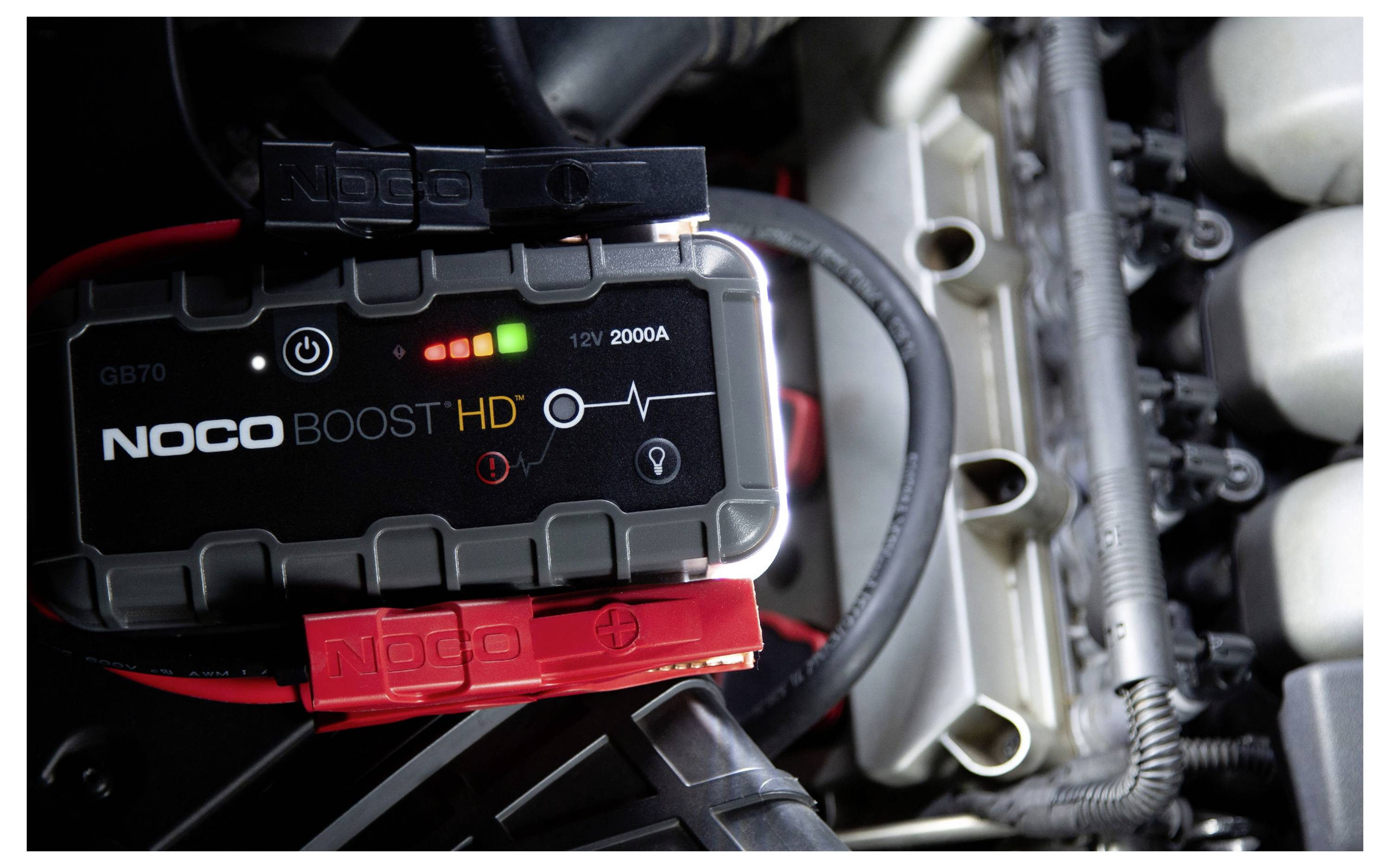 Un avviatore di emergenza NOCO Boost HD collegato nel vano motore di un'auto, che mostra le spie di carica della batteria e i cavi con pinze pronti per avviare il veicolo.