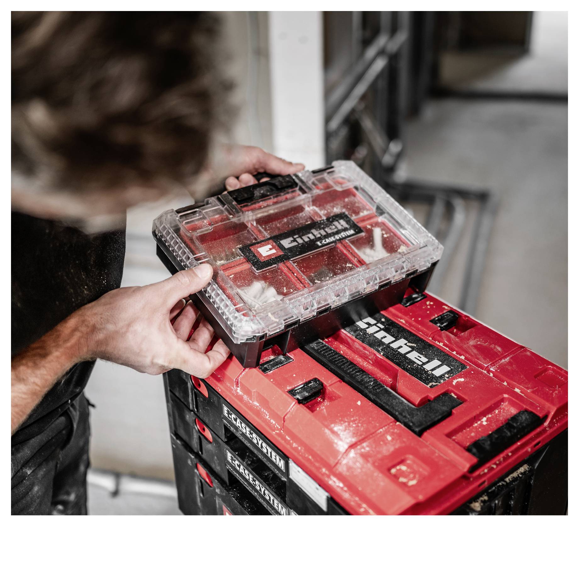 Una persona posiziona una cassetta per attrezzi Einhell con coperchio trasparente su un carrello portautensili rosso impilabile in un laboratorio o garage.