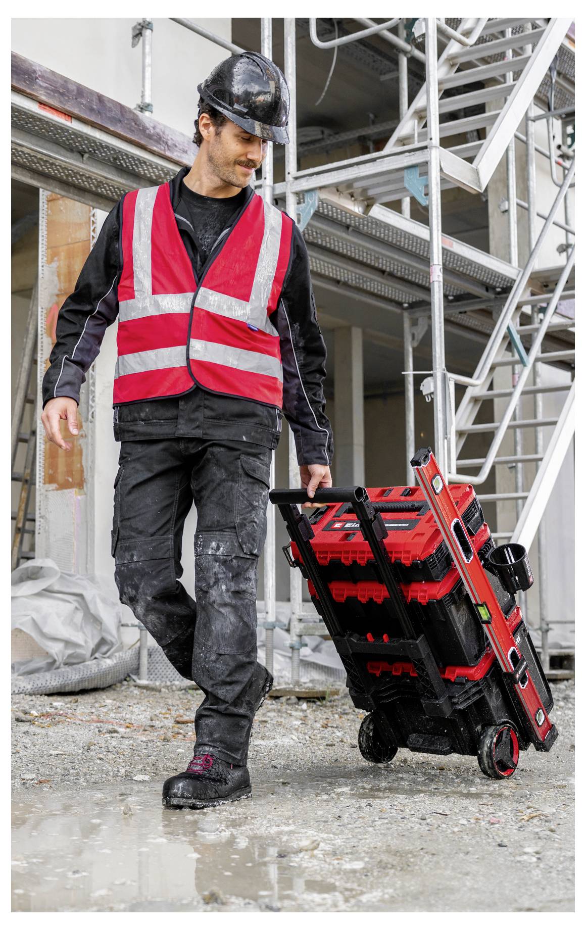 Un operaio edile con un casco di protezione e un gilet riflettente che tira un'attrezzatura su ruote in un cantiere.