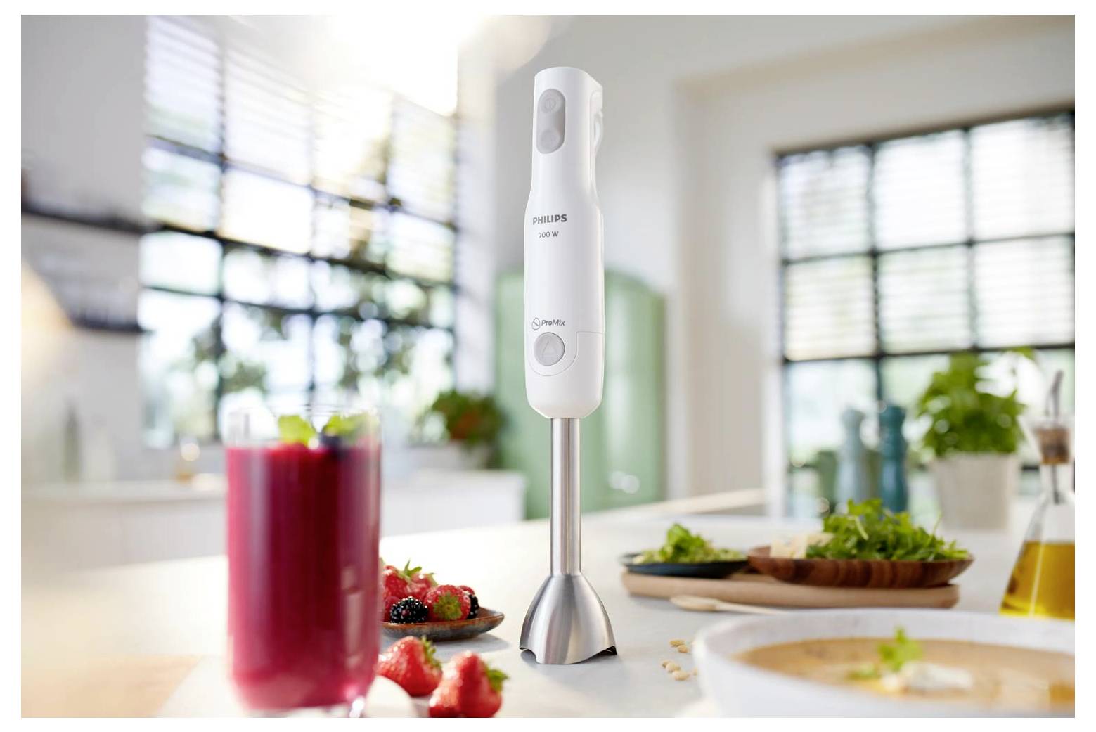 Frullatore ad immersione Philips in piedi su un piano di lavoro della cucina accanto a un frullato rosso e ingredienti preparati in una cucina domestica luminosa.