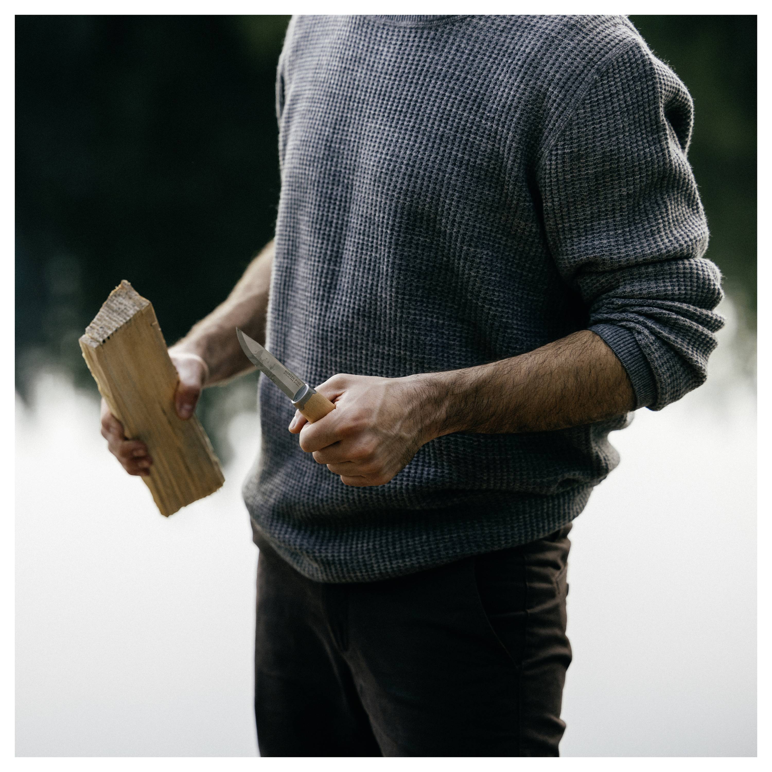 Persona in una felpa grigia che tiene un pezzo di legno e un coltello, pronta a intagliare o scolpire all'aperto.