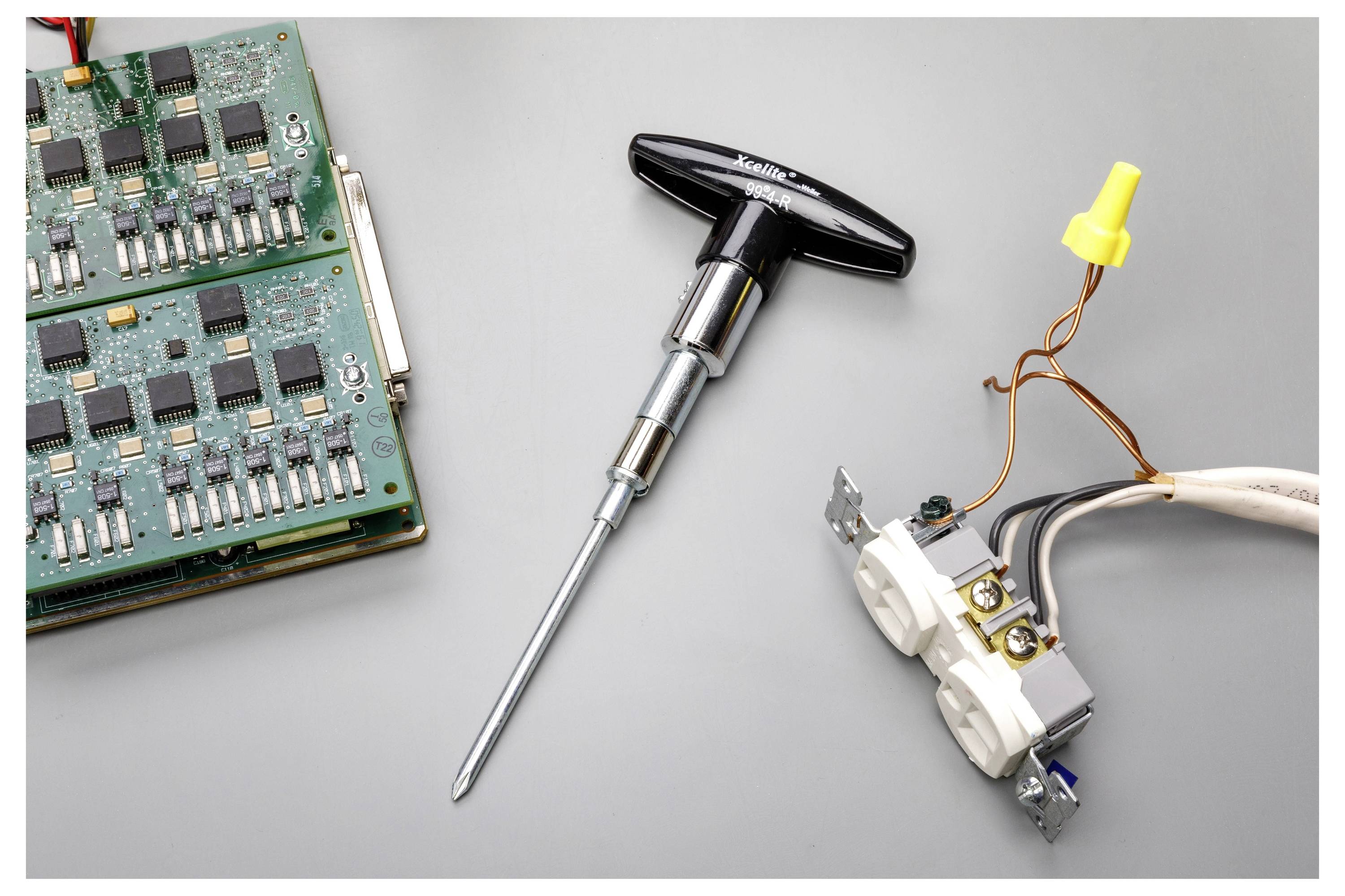 Scheda di circuito elettronico, cacciavite e parti di presa elettrica con cablaggio esposto su una superficie grigia, che suggerisce lavori di riparazione o montaggio.