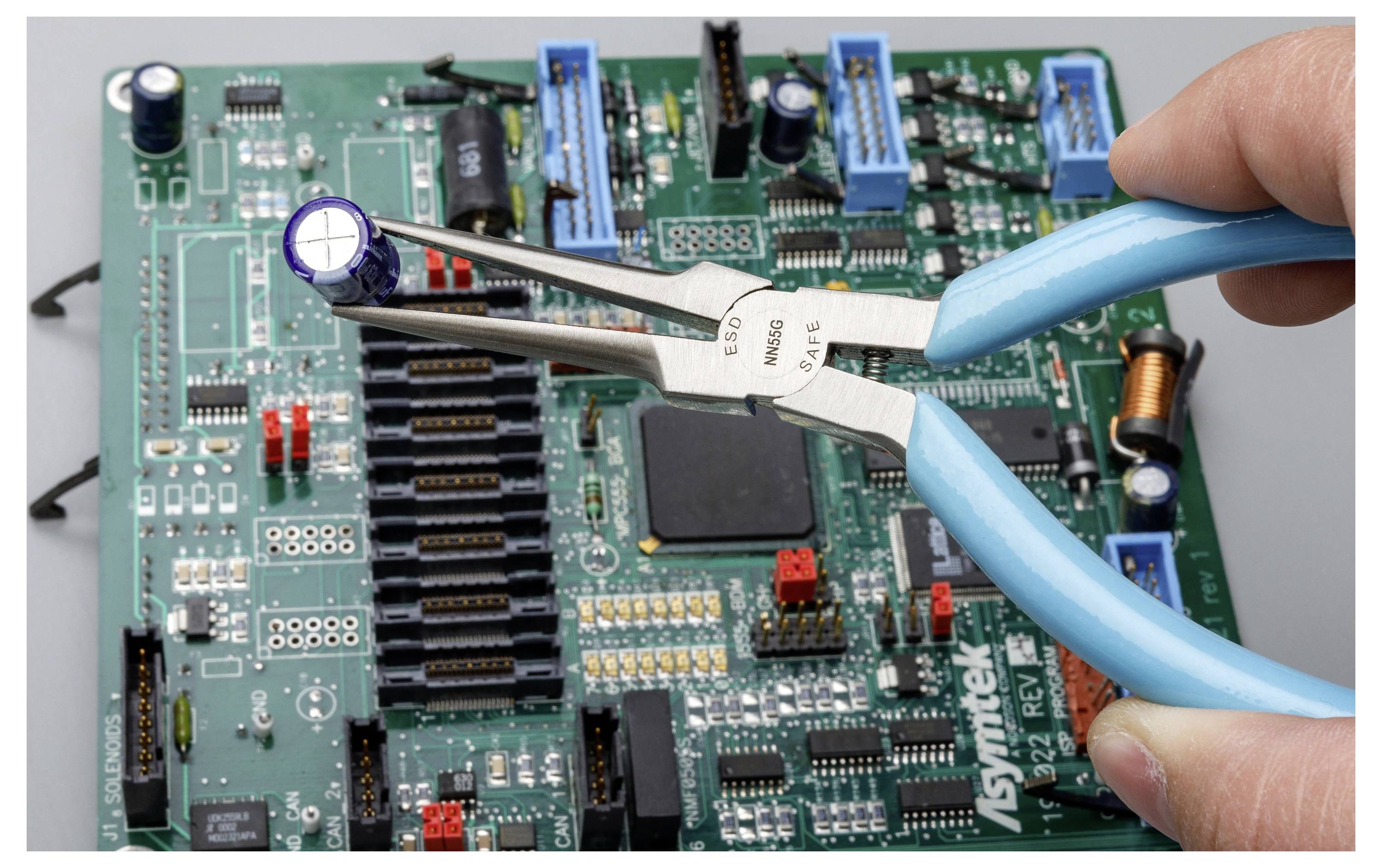 Una mano che tiene pinze blu rimuove un condensatore da una scheda di circuito, suggerendo una riparazione elettronica o l'estrazione di un componente.
