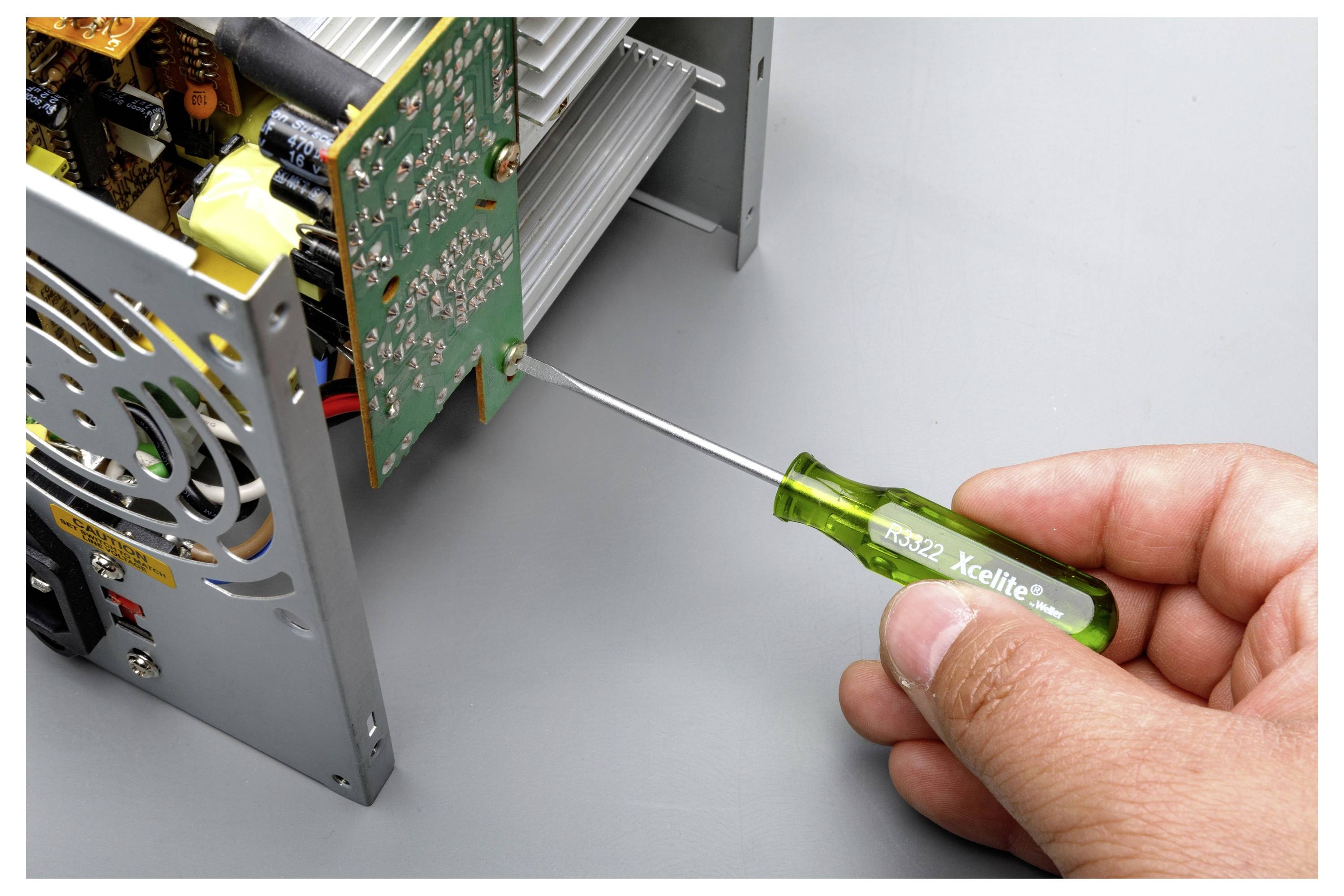 La mano utilizza un cacciavite Xcellite verde per serrare o rimuovere una vite sul lato di un dispositivo elettronico con scheda circuitale esposta.