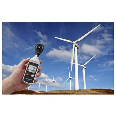 Mano che tiene un anemometro per misurare la velocità del vento di fronte a un parco eolico con diversi aerogeneratori sotto un cielo blu.