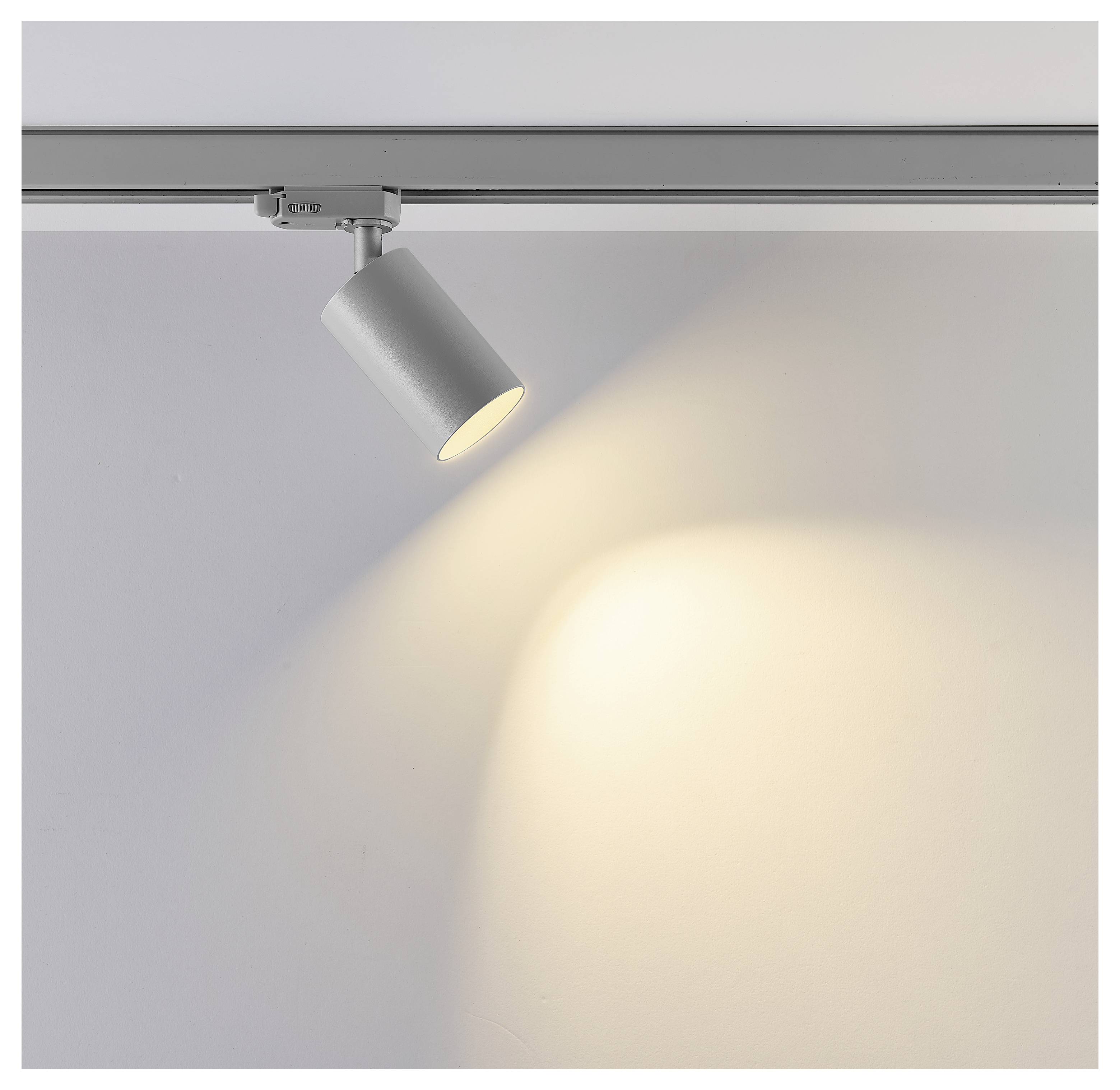 Apparecchio di illuminazione a binario in argento su rotaia a soffitto, orientato verso il basso e che proietta una calda luce riflettore su una parete vuota.