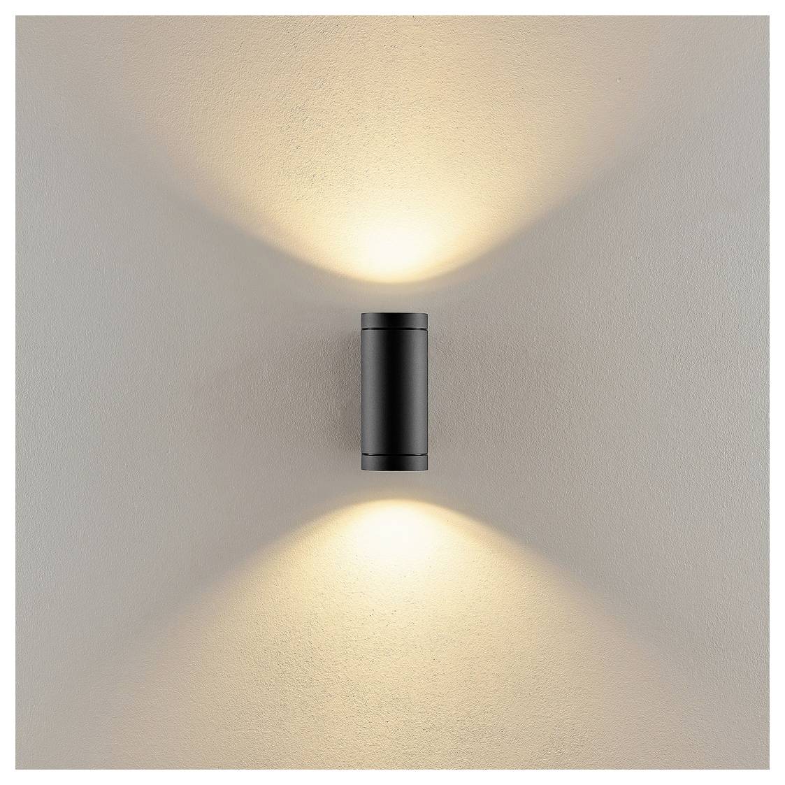 Applique murale cilindrica nera che diffonde una luce calda verso l'alto e verso il basso su una parete beige.