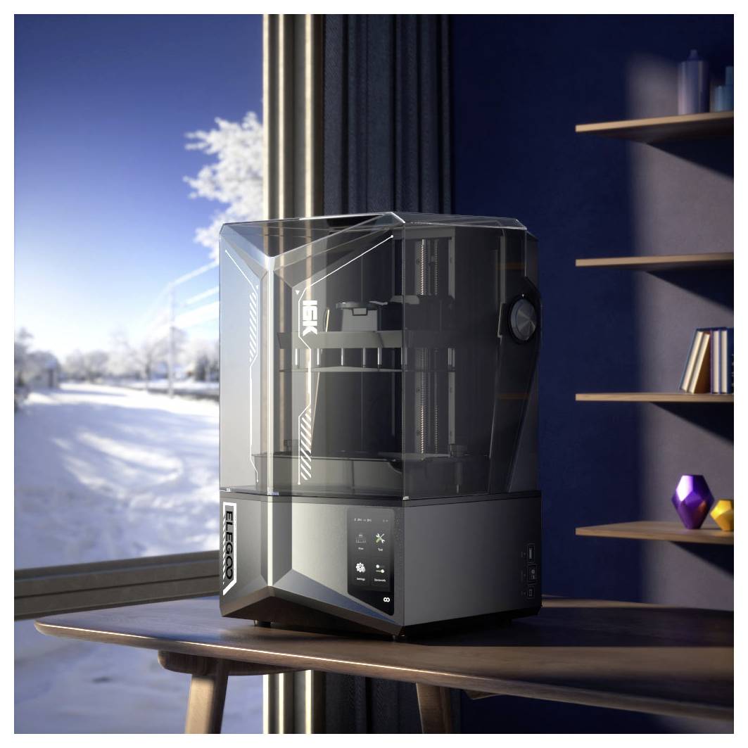 Una stampante 3D trasparente chiusa è posizionata su un tavolo vicino a una finestra, con scaffali sullo sfondo e un paesaggio innevato all'esterno.