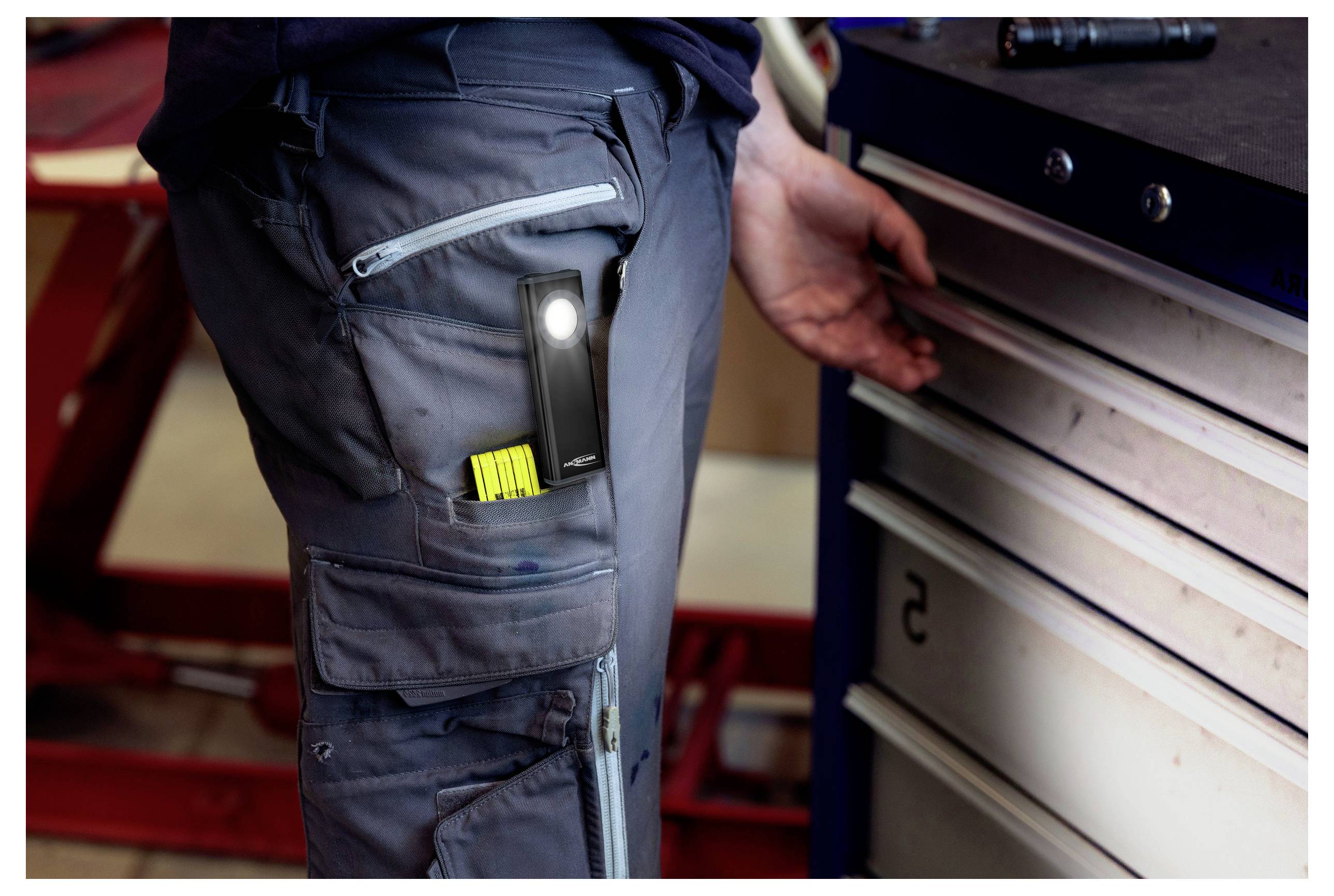 Primo piano dei pantaloni cargo di un operaio con una torcia elettrica agganciata e un pennarello giallo in tasca accanto a un cassettone per attrezzi in metallo.