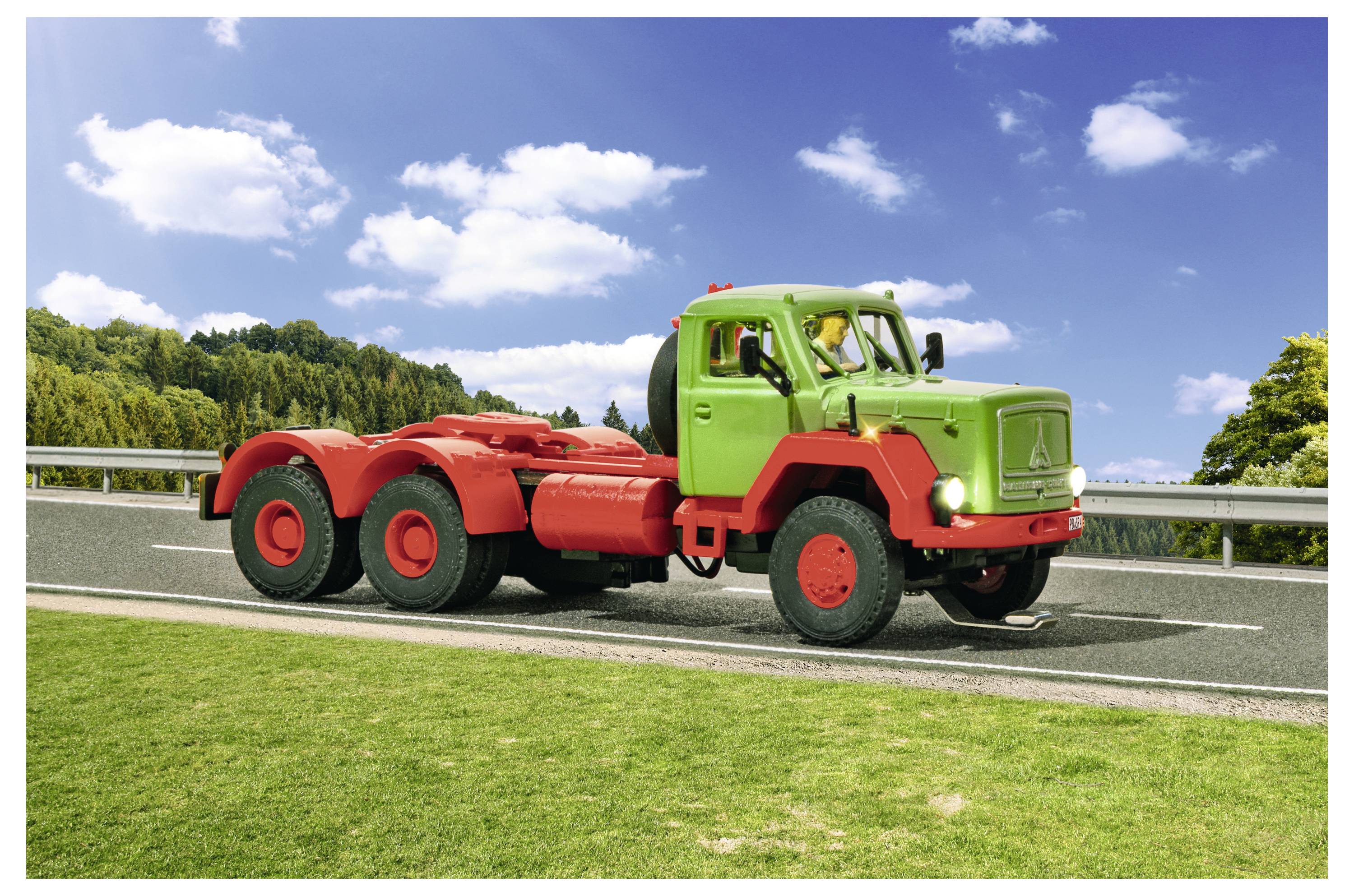 Camion a sei ruote di colore verde brillante e rosso parcheggiato sul margine di una strada con erba in primo piano, alberi e cielo blu con nuvole.