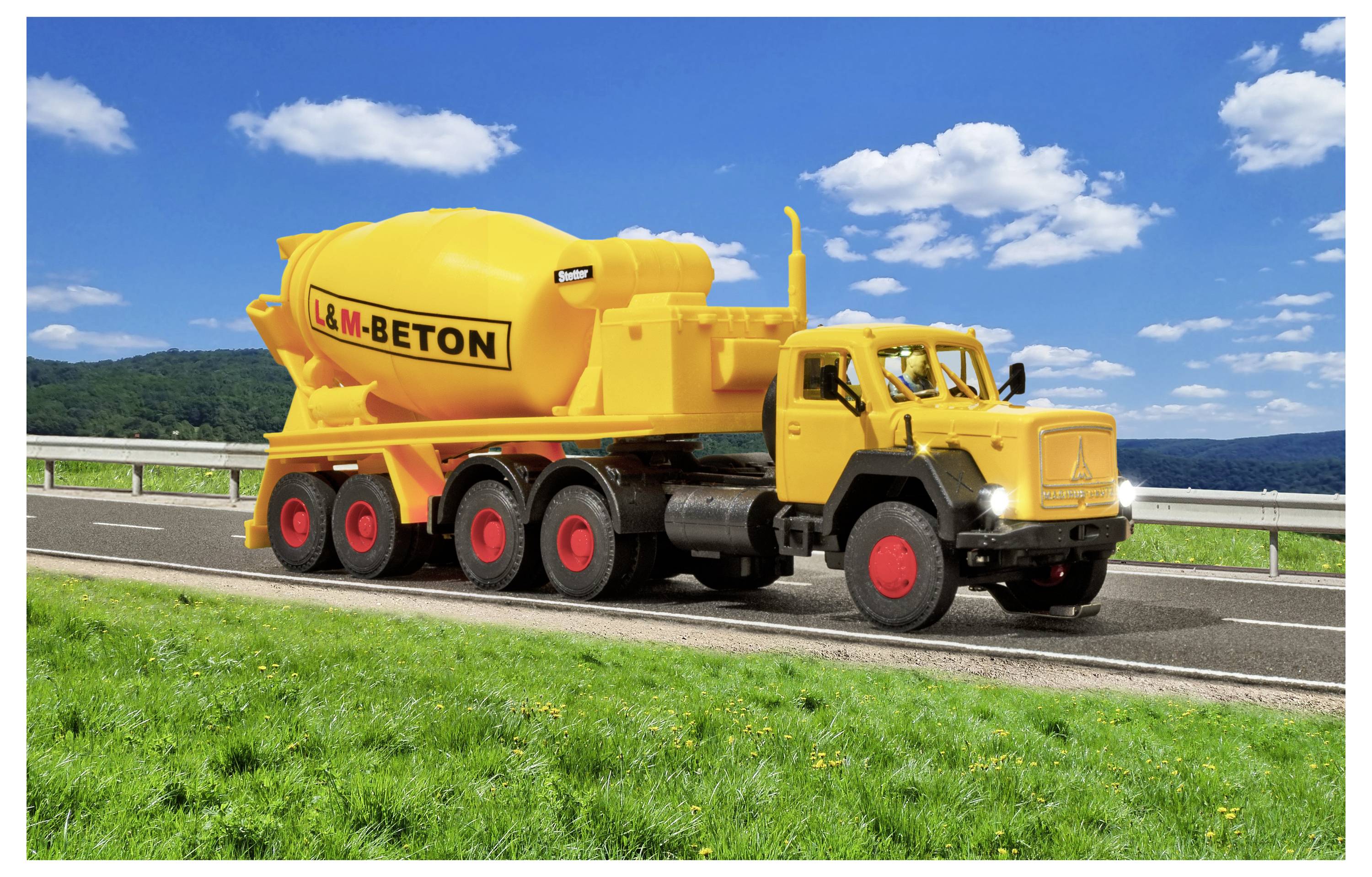 Camion betoniera giallo 'L&M-BETON' parcheggiato su un ciglio della strada accanto all'erba verde sotto un cielo blu con nuvole.