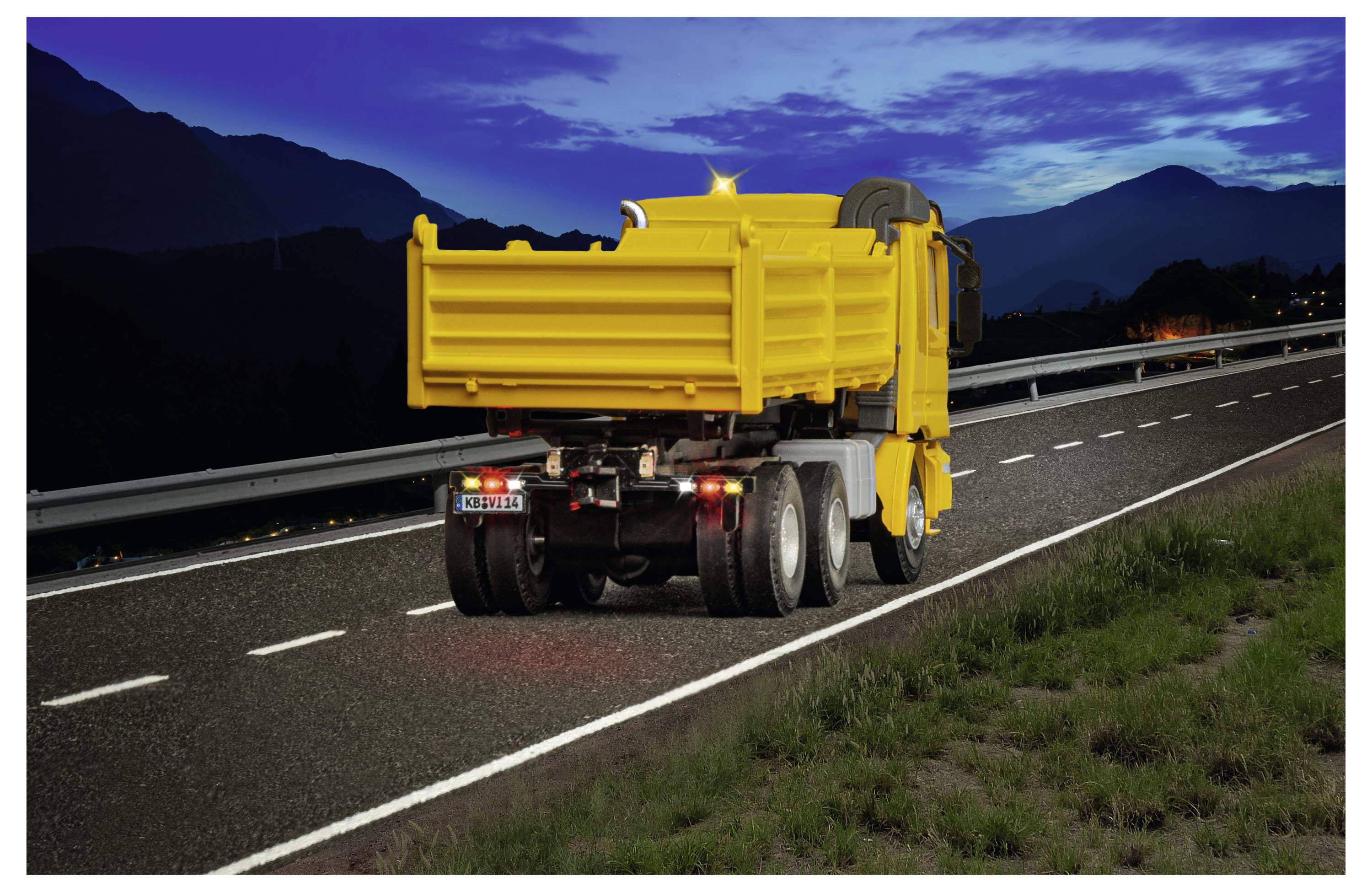 Camion ribaltabile giallo che viaggia su una strada di montagna scura al tramonto, visto da dietro con i fari posteriori accesi.