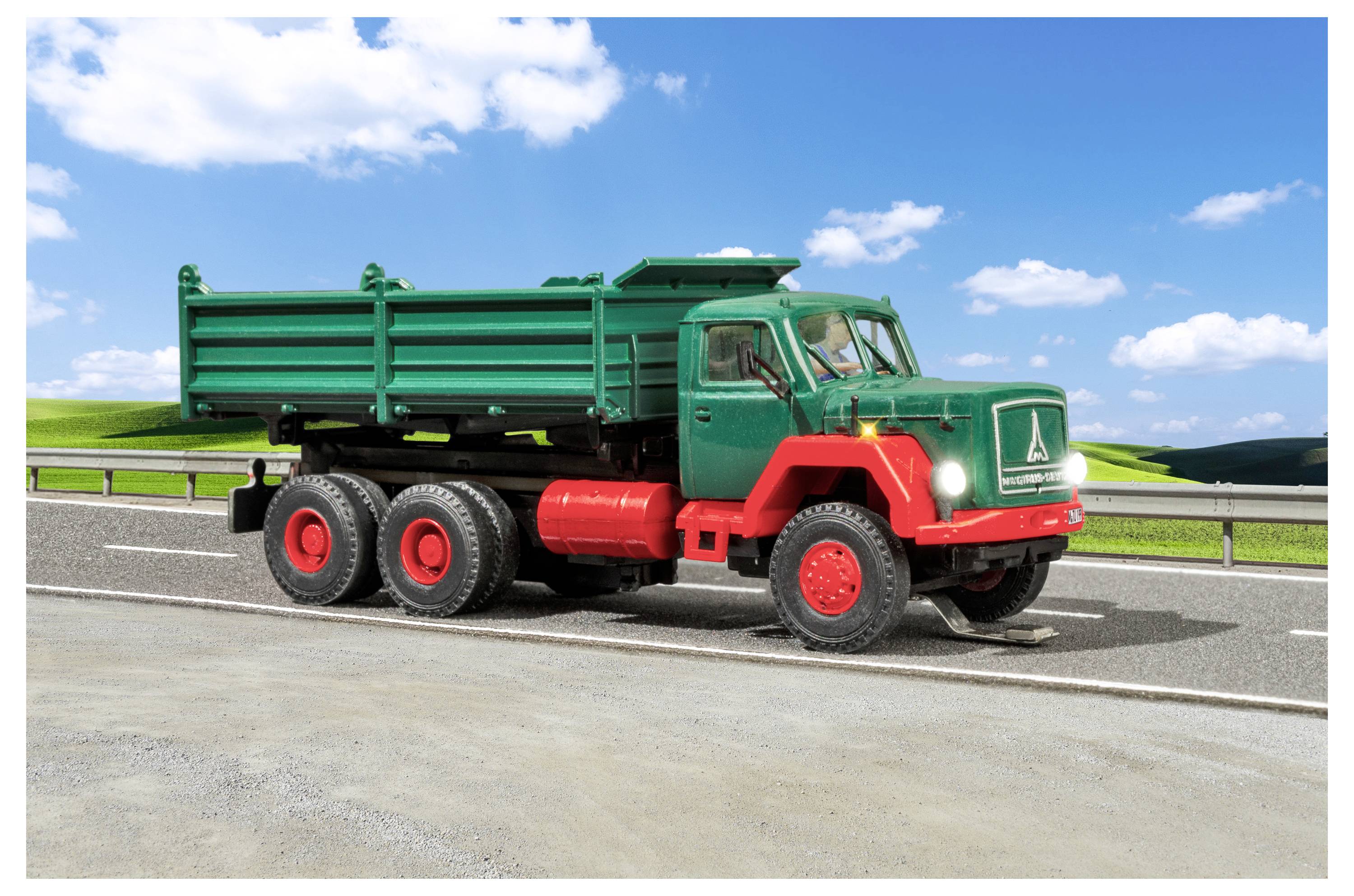 Camion ribaltabile verde a sei ruote che guida su una strada sotto un cielo blu luminoso con nuvole, mostrato in vista laterale.