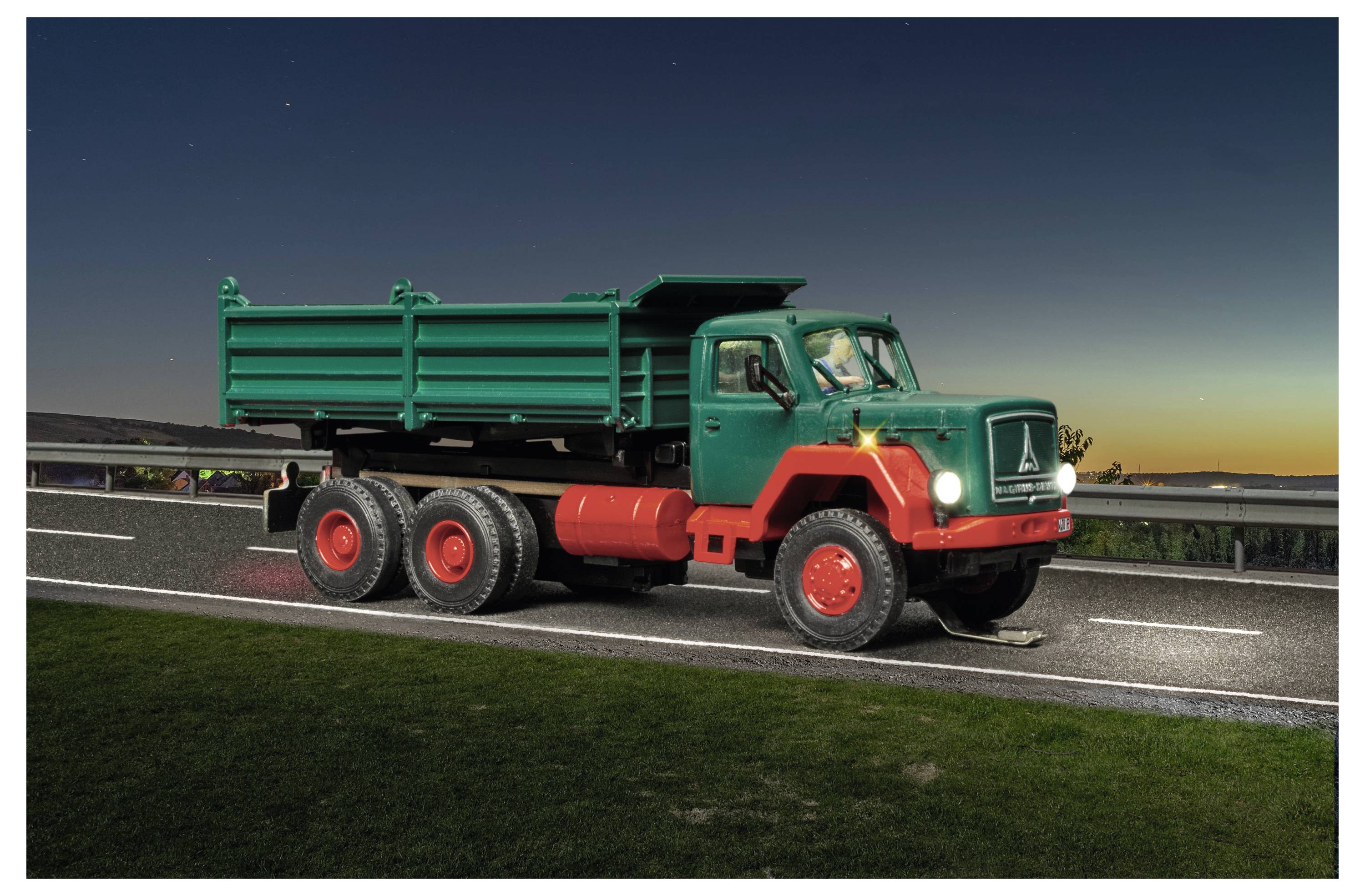 Camion ribaltabile verde con ruote rosse che guida su una strada al crepuscolo, visto di lato.