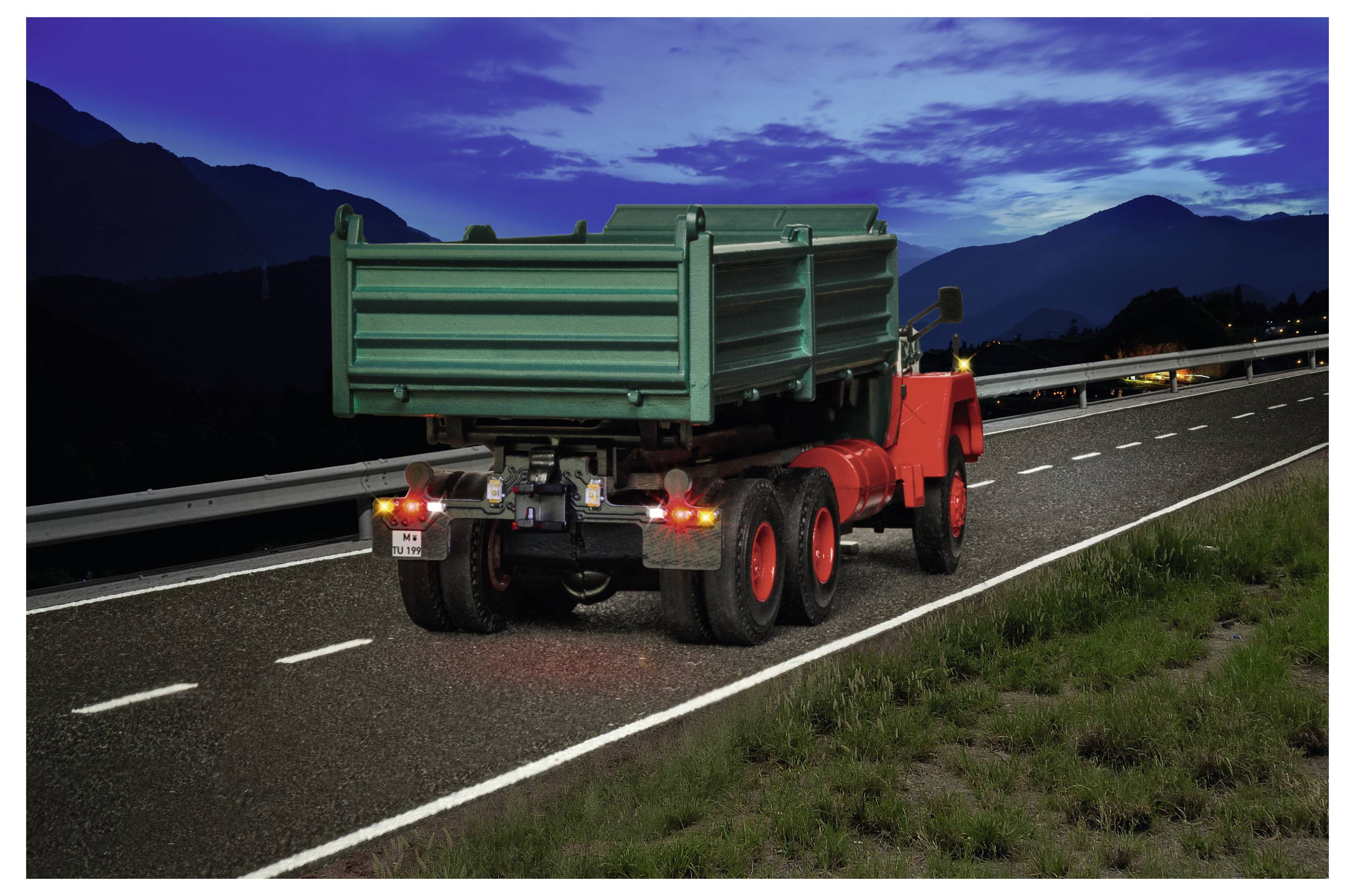 Camion ribaltabile rosso con cassone di carico verde sollevato che procede su un'autostrada al tramonto accanto a montagne e guardrail.