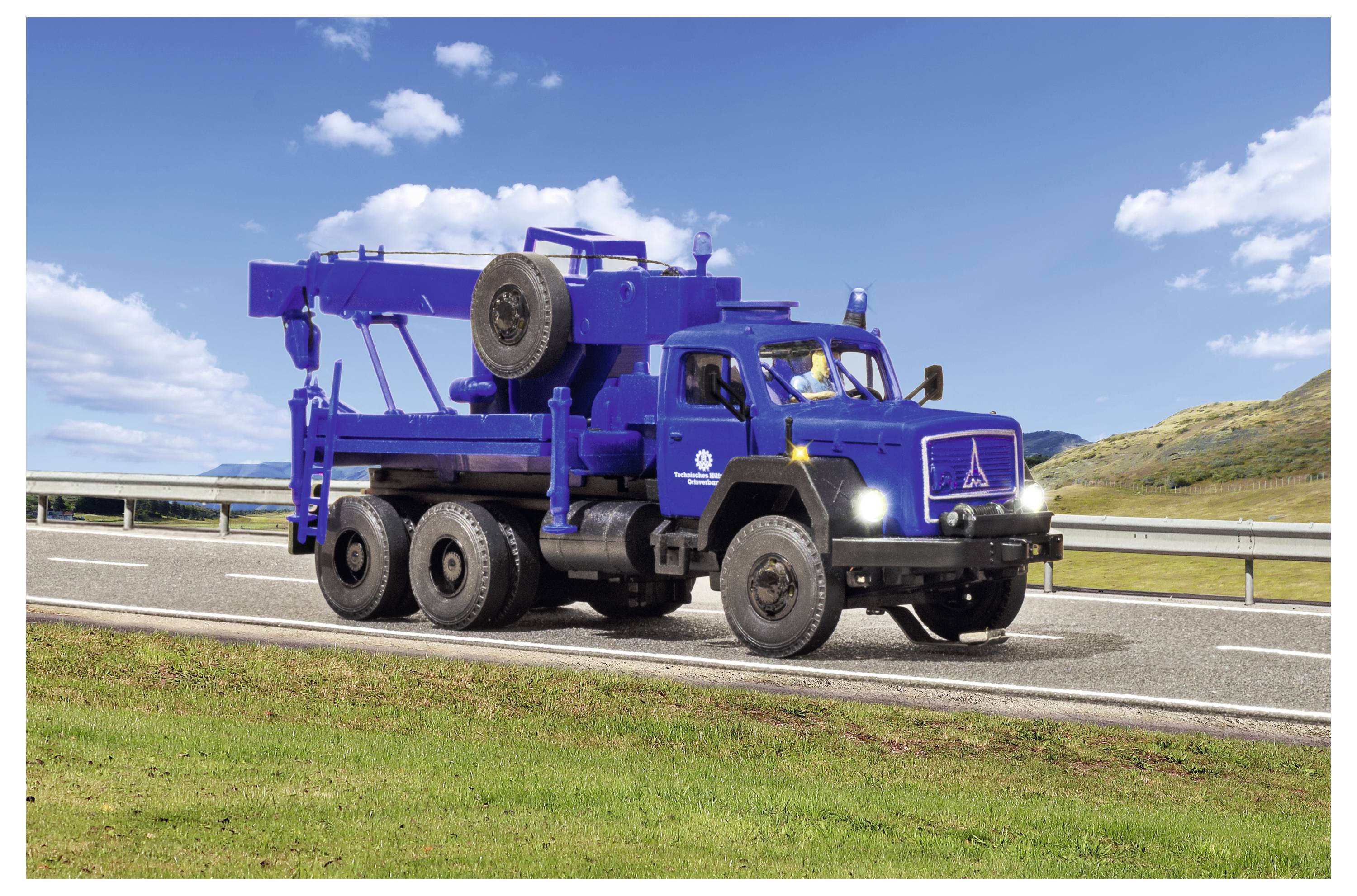 Camion di soccorso stradale blu pesante che guida in autostrada sotto un cielo azzurro, con una gru montata e una ruota di scorta sul retro.