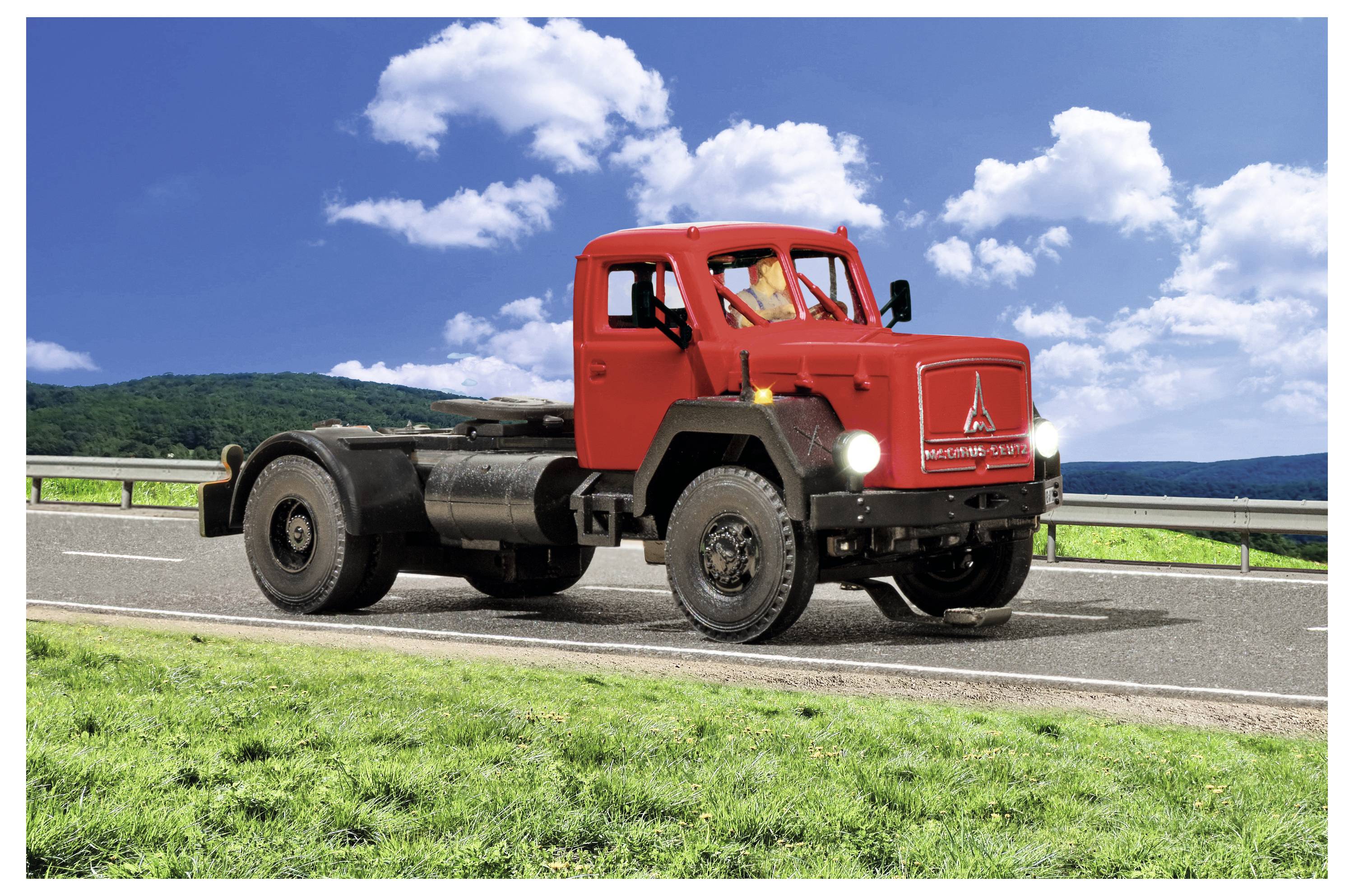 Cabina di un camion vintage rosso su telaio parcheggiato a bordo strada con erba verde, montagne e un cielo blu con nuvole sullo sfondo.