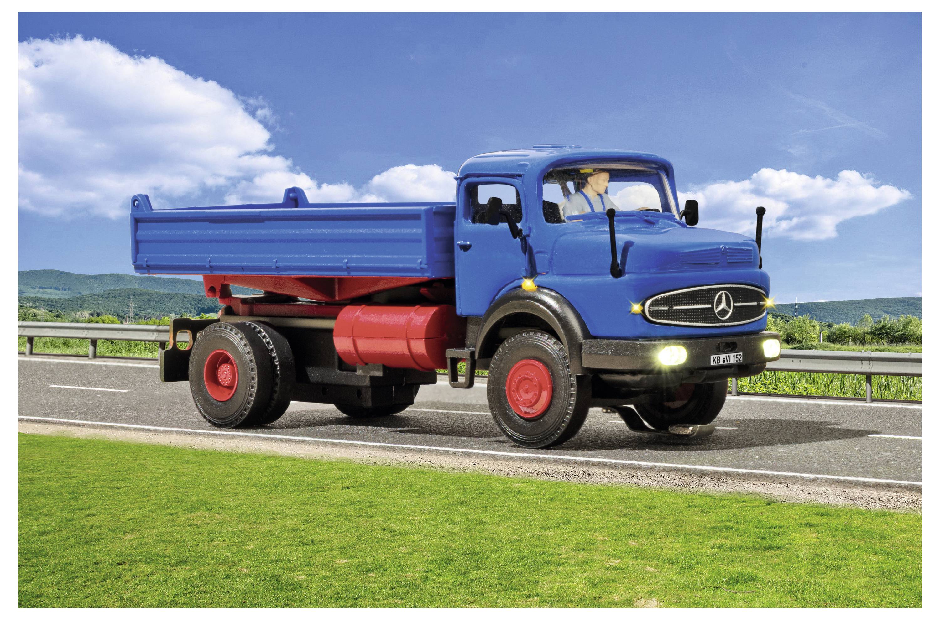Camion ribaltabile blu che guida su una strada accanto all'erba verde sotto un cielo parzialmente nuvoloso.