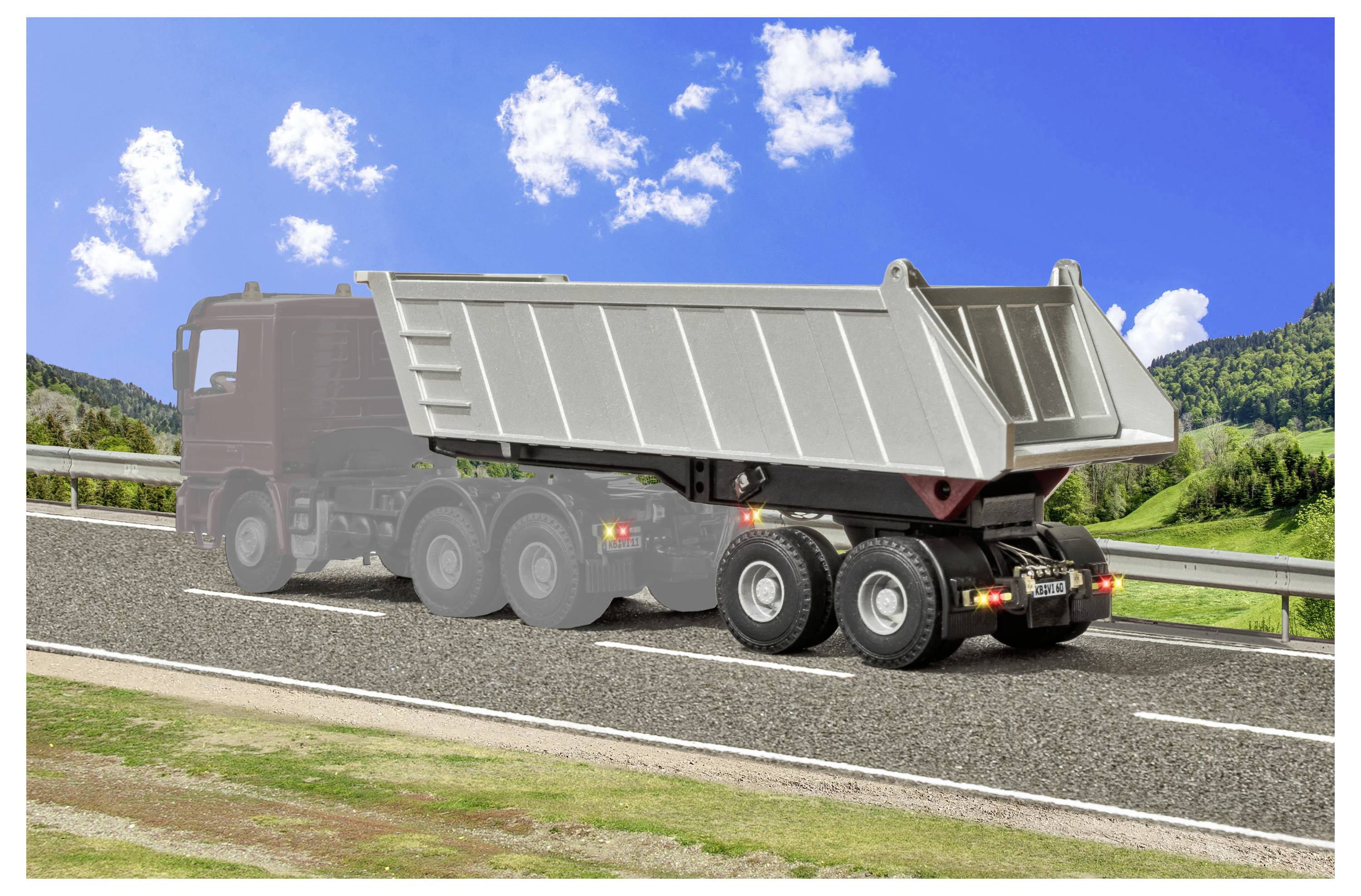 Camion ribaltabile grigio che guida su un'autostrada sotto un cielo blu con nuvole e colline verdi sullo sfondo.